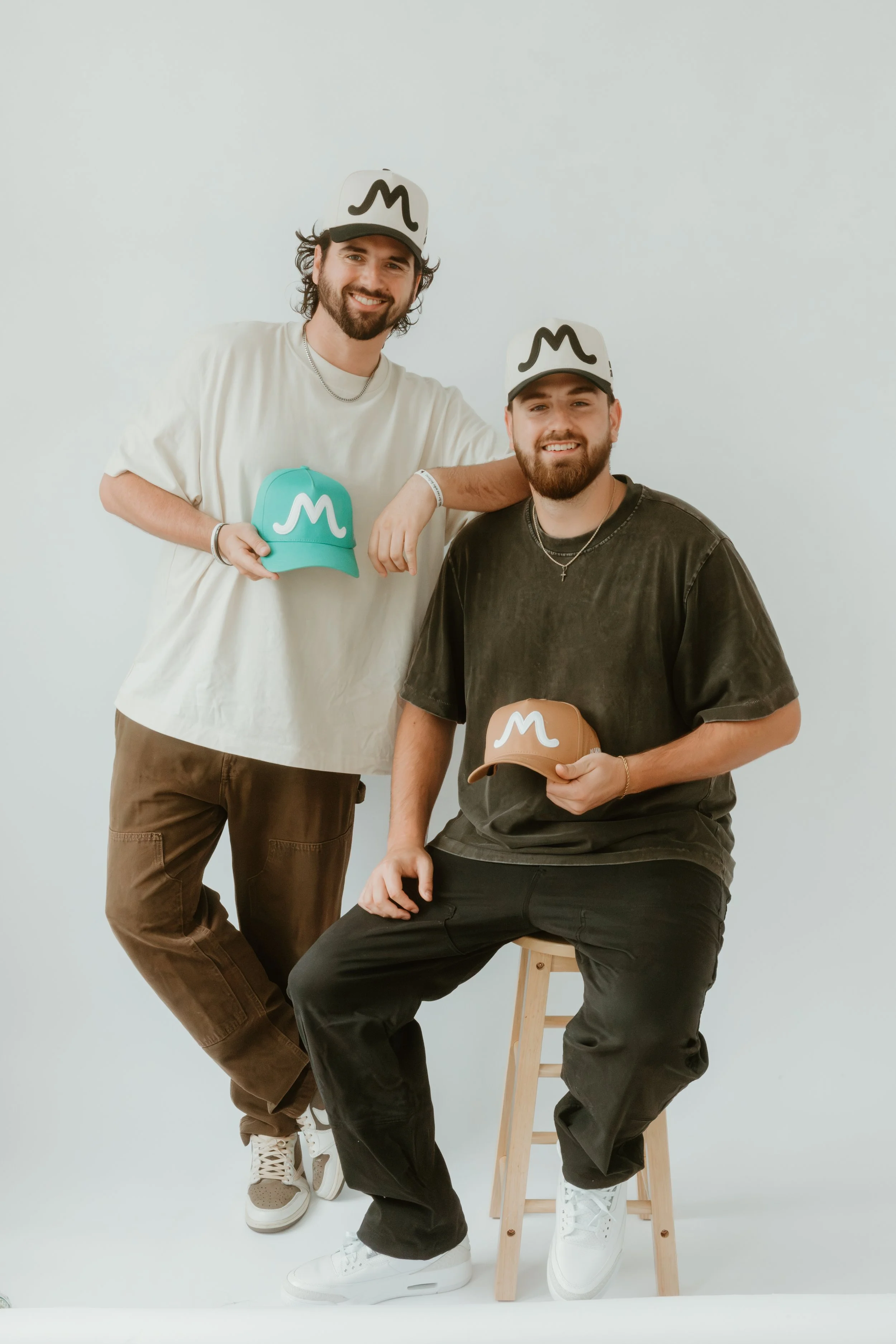 Two young men smiling, one standing with a hand on the other man's shoulder, both wearing white and black baseball caps with an M logo, holding additional caps, and wearing casual clothes against a plain white background.