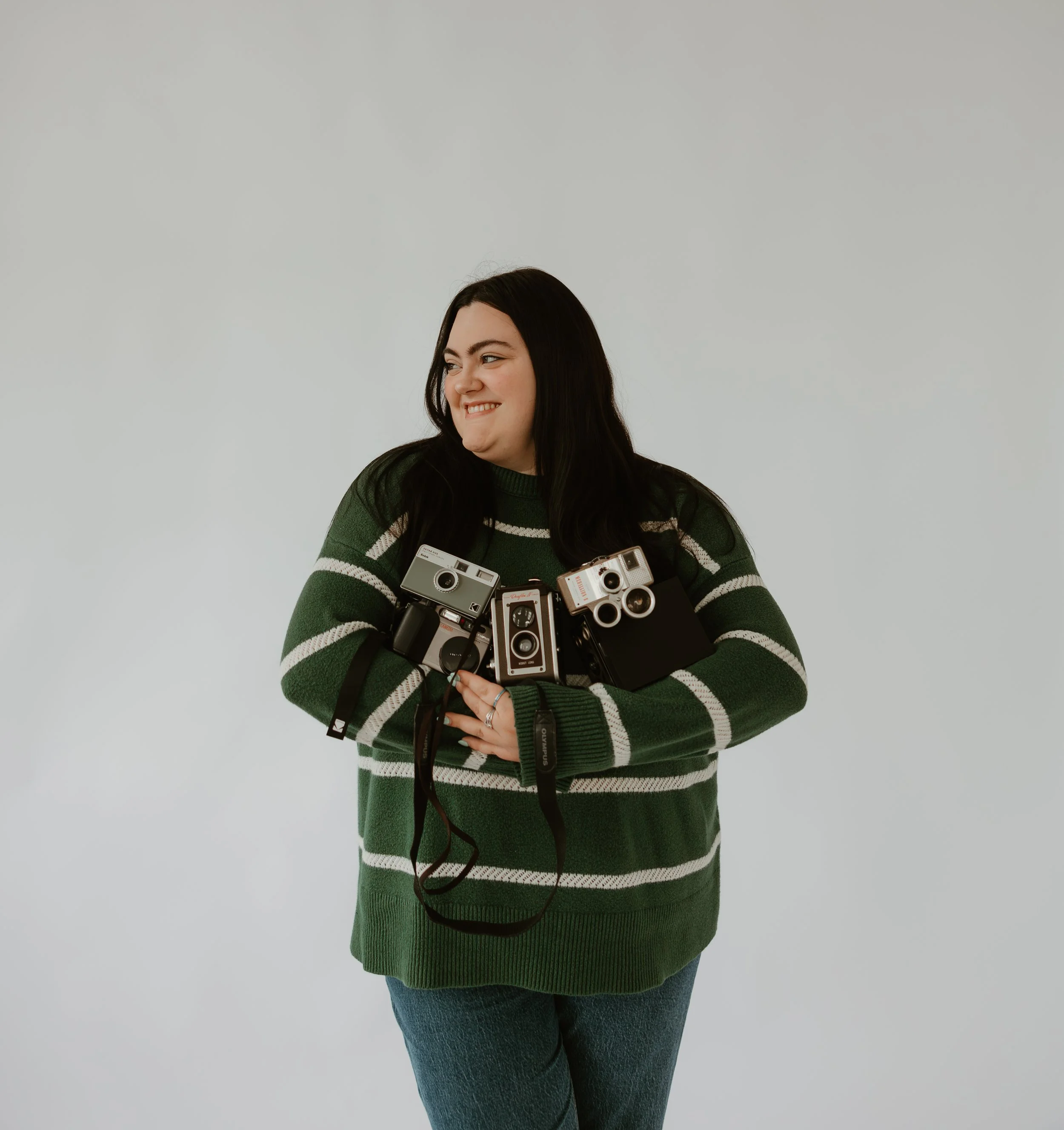 A woman wearing a green and white striped sweater and blue jeans, holding multiple cameras, standing against a plain light gray background.