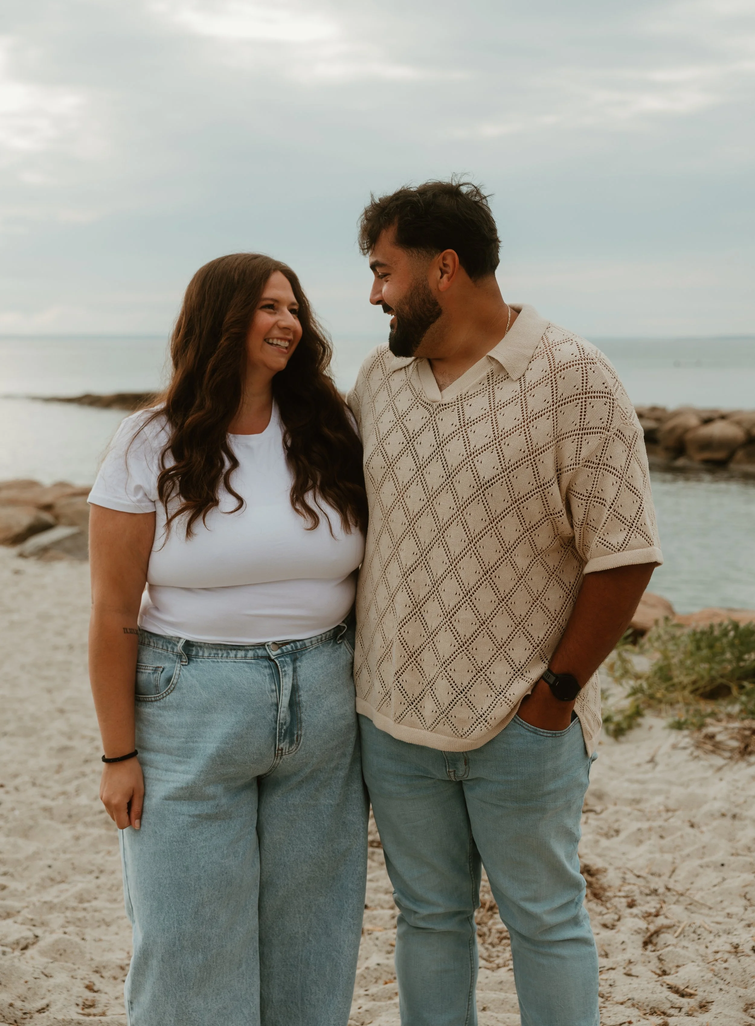 A smiling couple standing together on the beach, facing each other, with the ocean and rocks in the background.