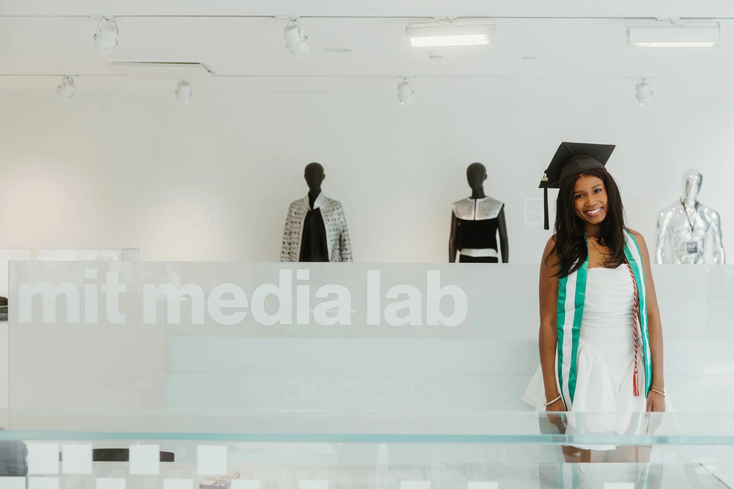 Young woman smiling in front of the MIT Media Lab reception desk, wearing a graduation cap and gown with green and white stripes, with mannequins dressed in fashion behind her.