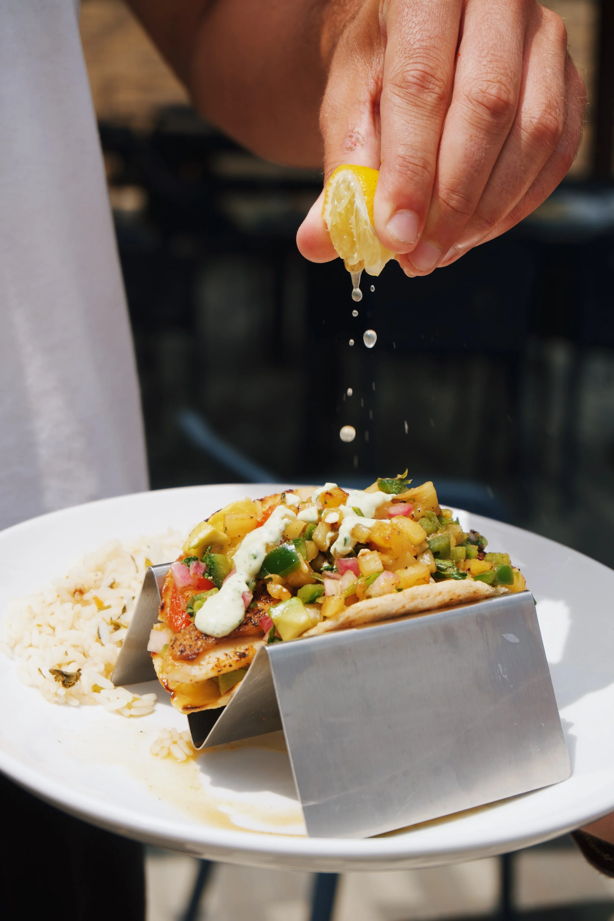 Person squeezing lemon juice onto a taco with diced vegetables and sauce, served on a white plate.