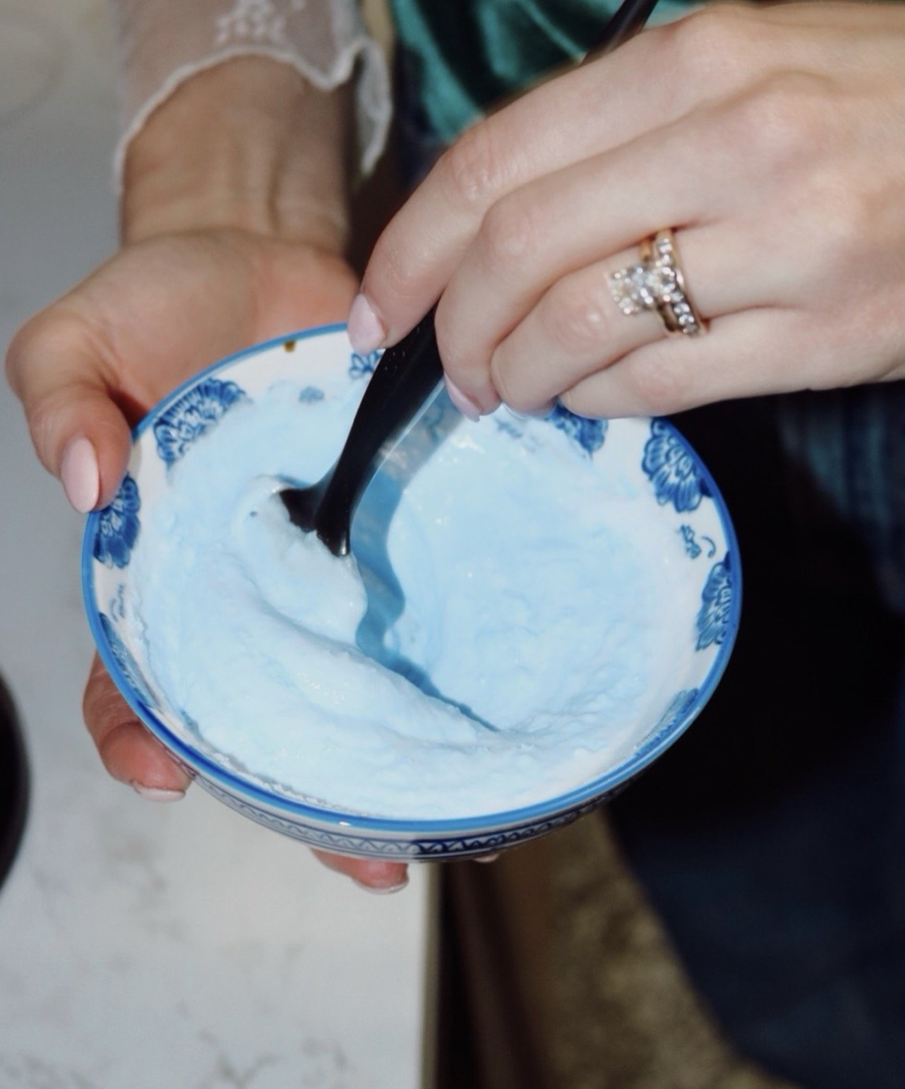 Person stirring a light blue frozen dessert or ice cream in a blue and white patterned bowl with a black spoon, wearing a ring with multiple stones.