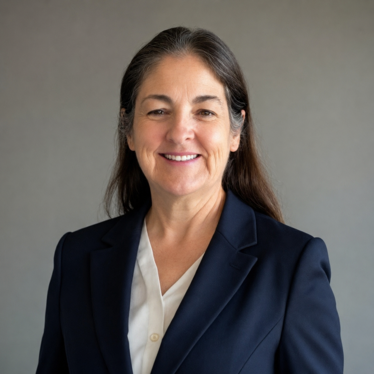 Professional woman with long brown hair in a navy blazer and white blouse smiling at camera against a neutral gray background.