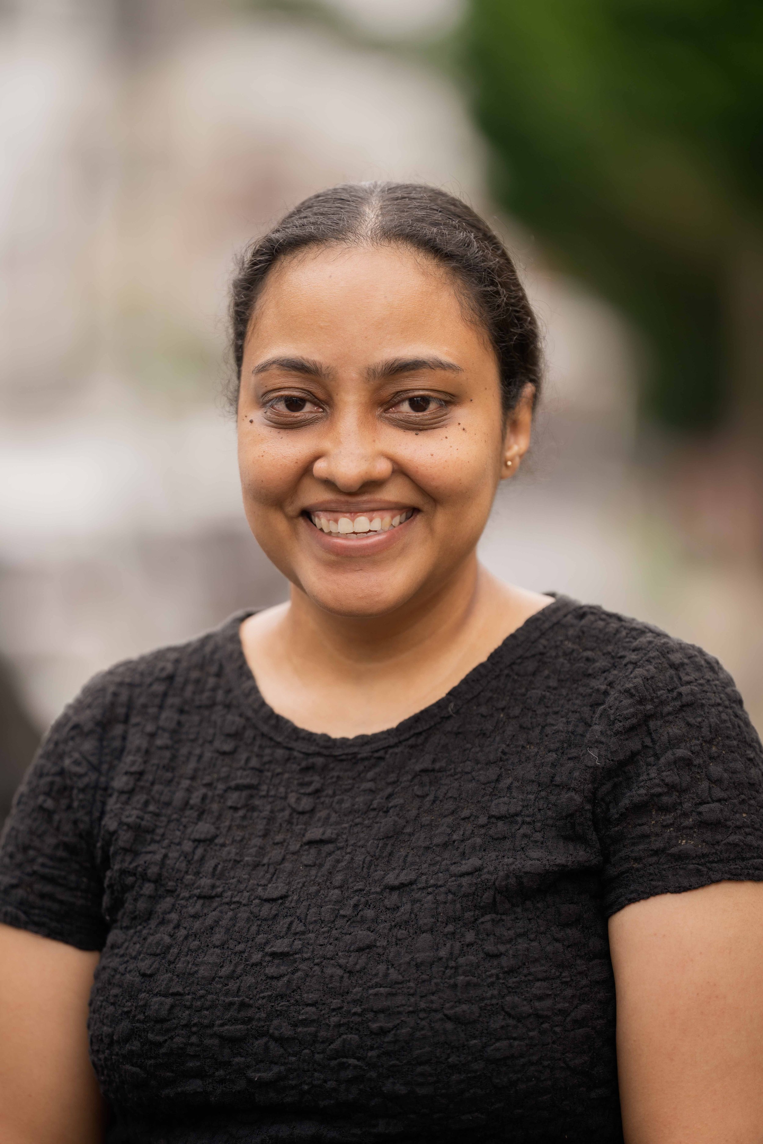 portrait of a smiling woman with dark hair pulled back, wearing a black textured top, standing outdoors with blurred background.