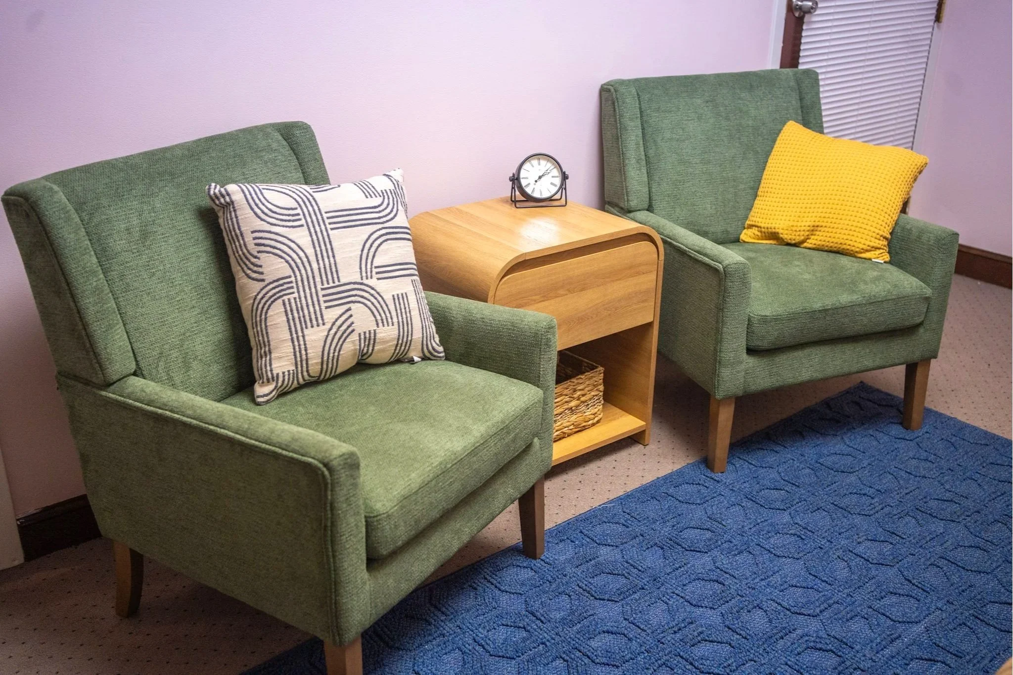 Two green upholstered armchairs with wooden legs, each with a decorative pillow, are placed on a beige carpet next to a small wooden table with a black clock on it. The wall behind is plain and light-colored, and there is a door with blinds partially visible on the right.