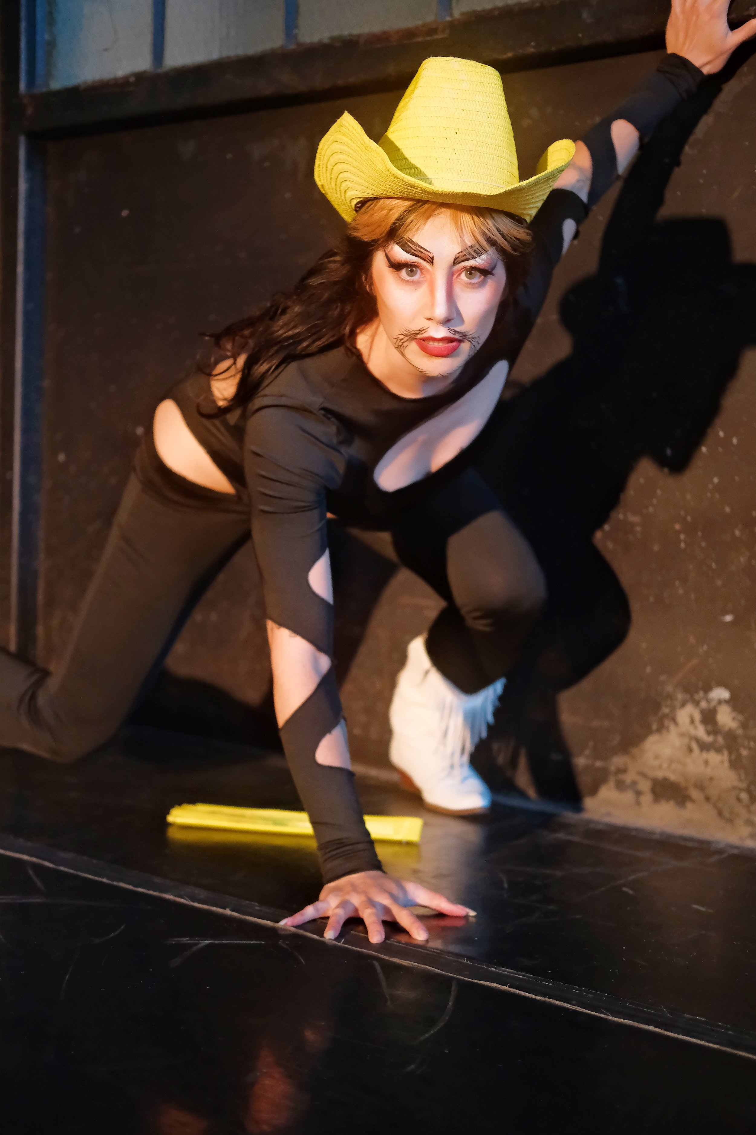 Deuce Lee - a Chinese American drag king with thick black calligraphic eyebrows and goatee, perches in a runner stance, wearing a black cutout bodysuit, white fringe cowboy boots and a yellow cowboy hat
