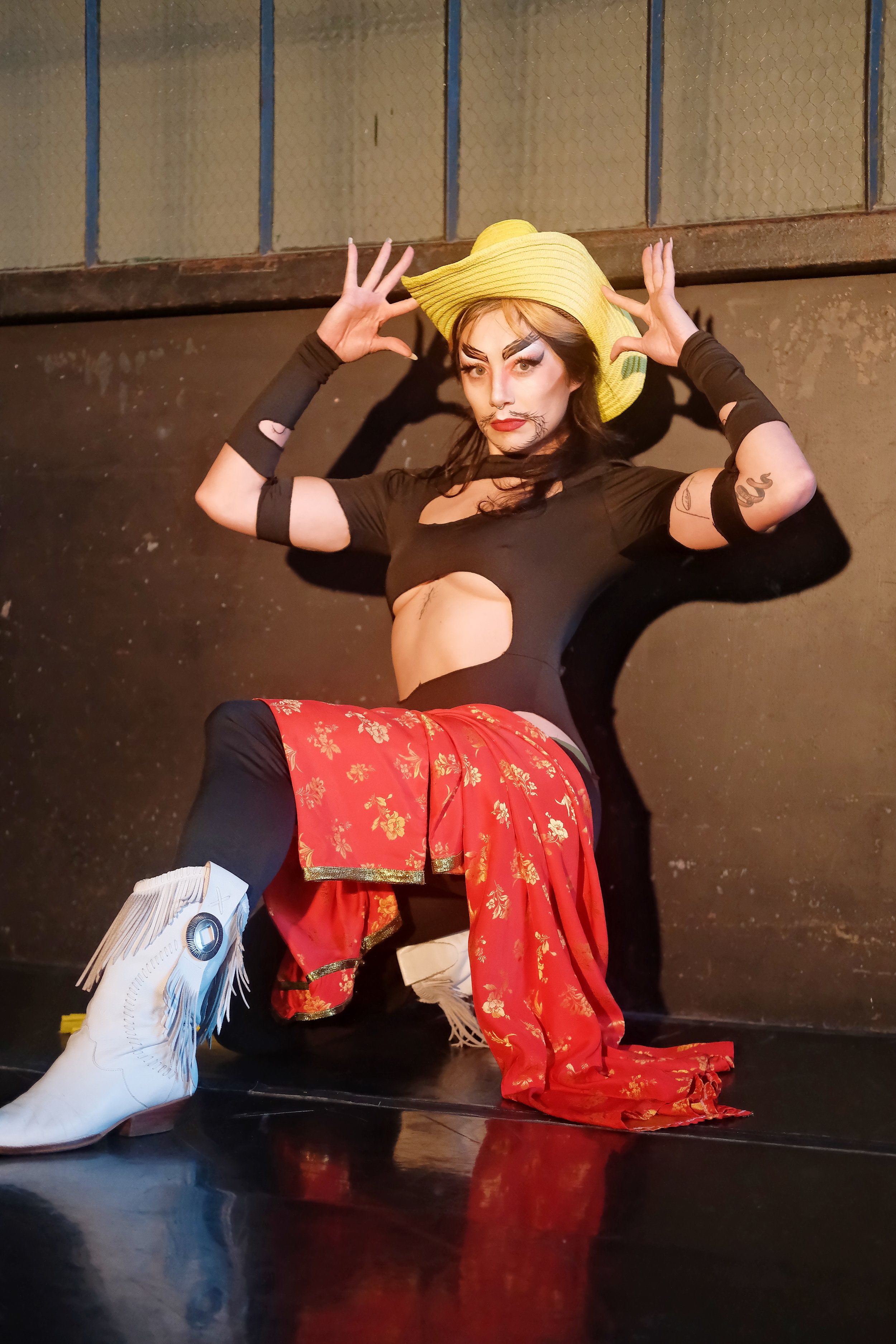 Deuce Lee - a Chinese American drag king with thick black calligraphic eyebrows and goatee, kneeling with their hands on their hat, wearing a black cutout bodysuit, white fringe cowboy boots and a yellow cowboy hat