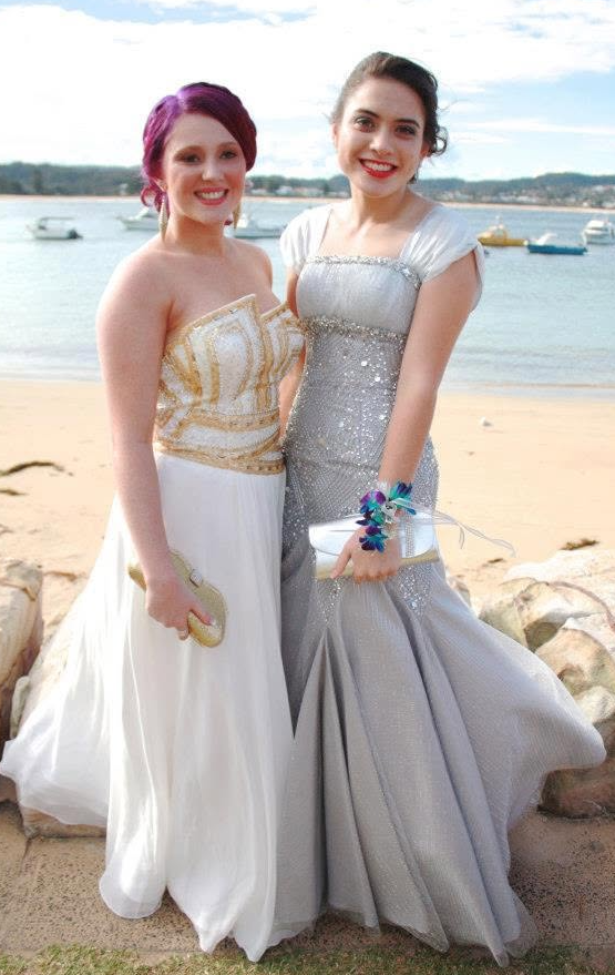 Two women in formal dresses standing on a beach with boats in the water behind them.