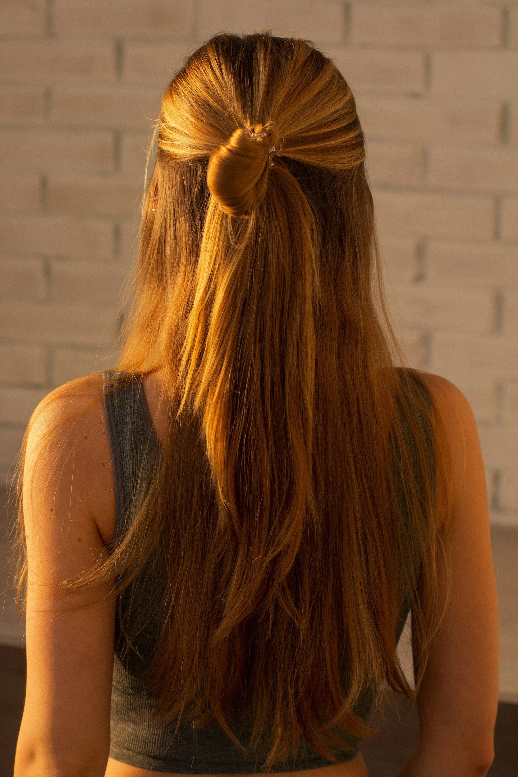 Back of a woman with long, reddish hair styled in a half-up bun, standing against a light-colored brick wall.