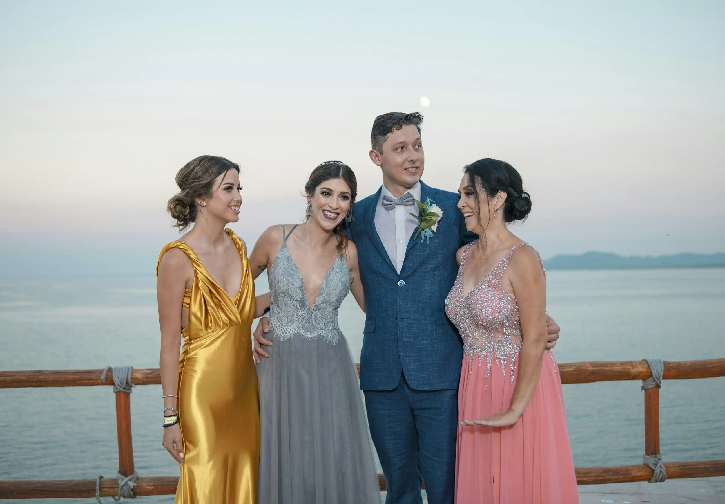 Four people in formal attire standing on a boat railing with water and mountains in the background, smiling and chatting during sunset.