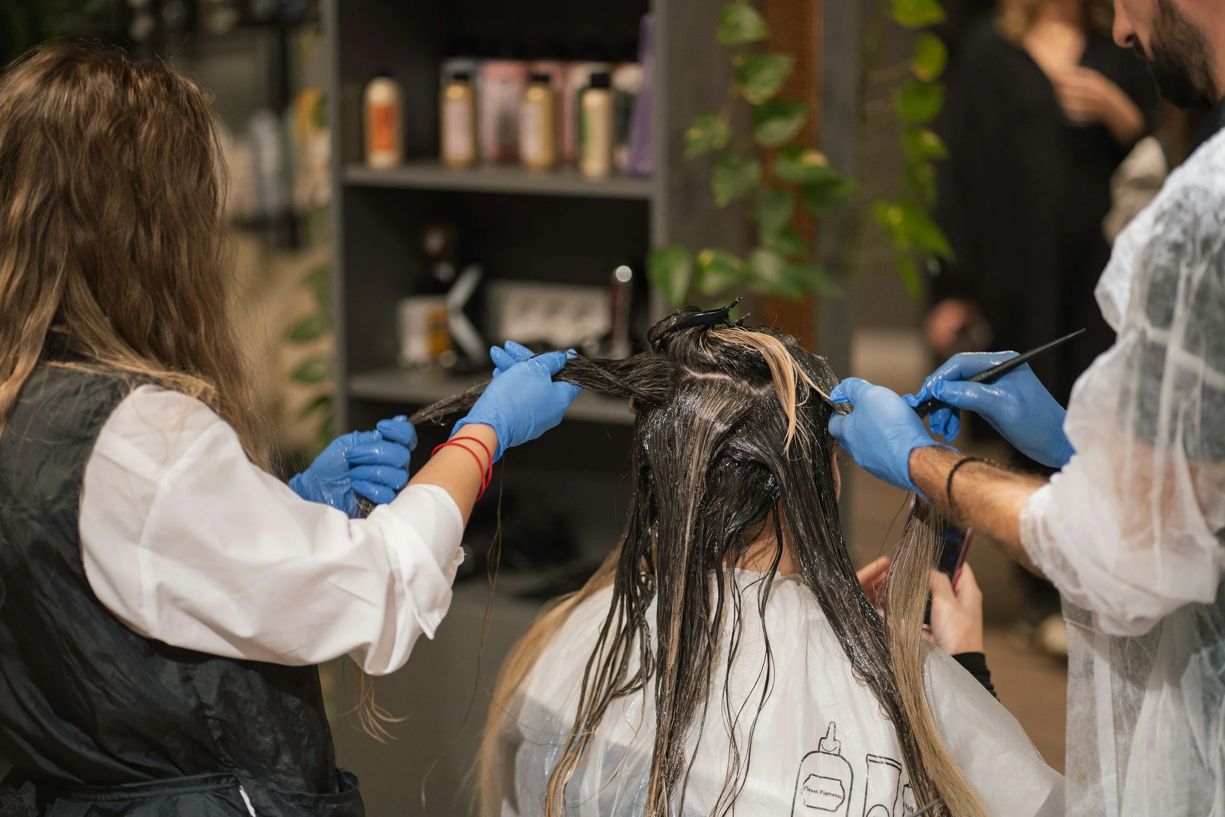 Hair salon or barber shop where a stylist is applying hair dye or highlights to a client's hair, with another person assisting.