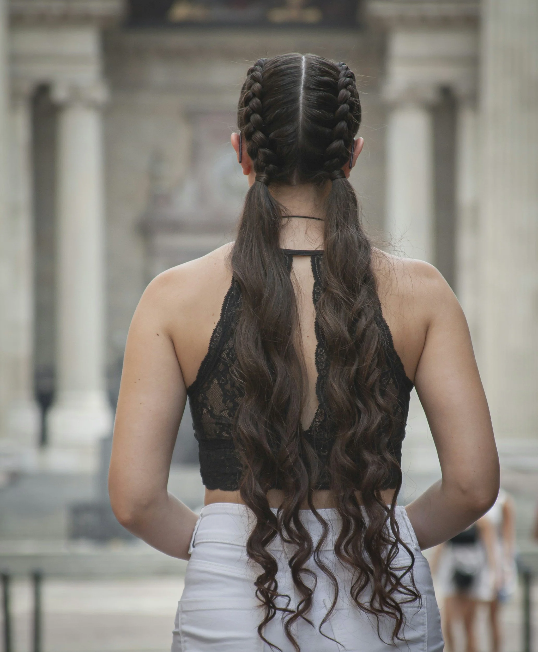 Back view of a woman with long, wavy brown hair styled in two French braids, wearing a black lace sleeveless top and white pants, standing outdoors near what appears to be a public place with columns and people in the background.