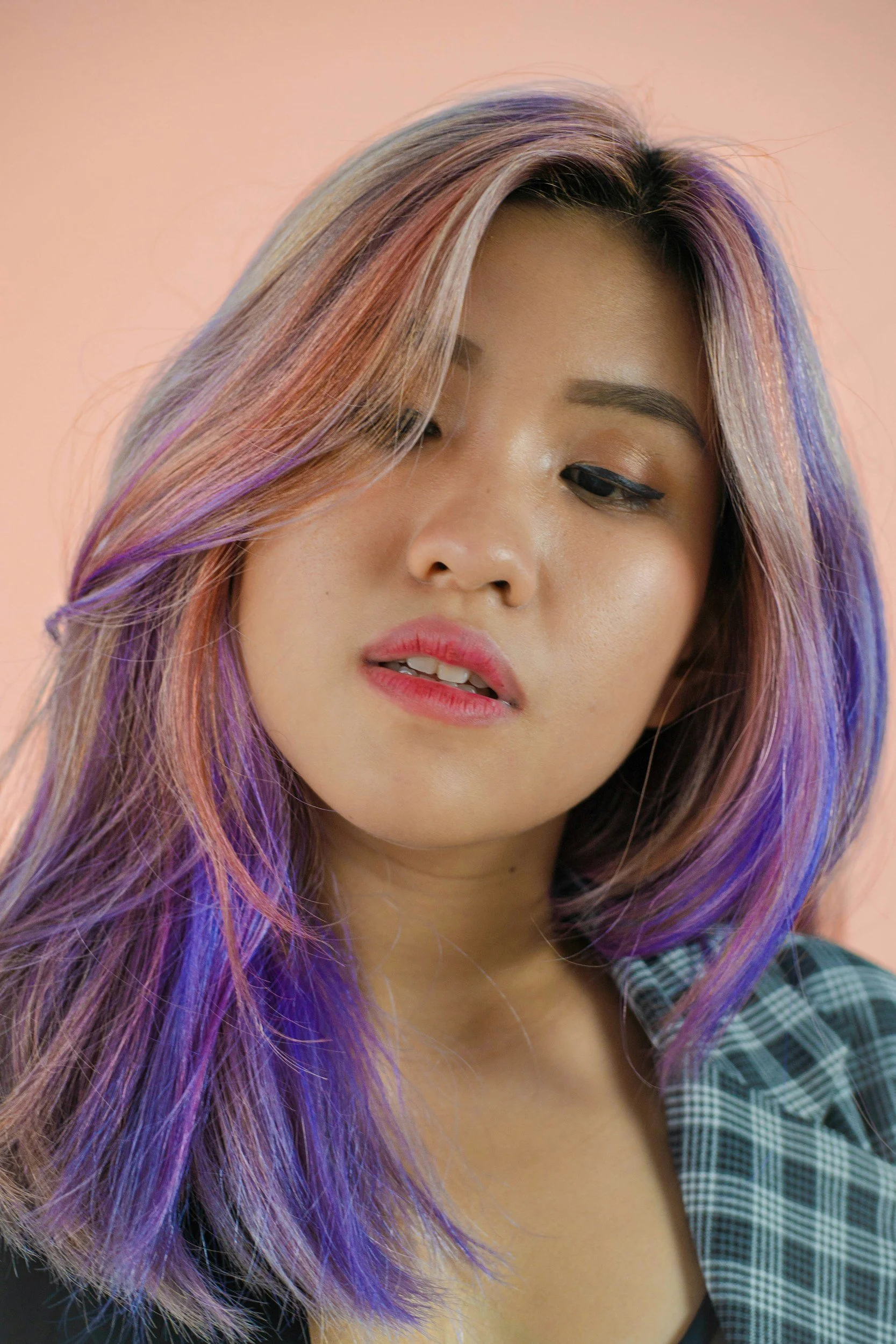 Close-up of a young woman with pastel-colored hair in shades of pink, purple, and blue, wearing a plaid shirt. Her face is gently turned, showing her makeup and relaxed expression.
