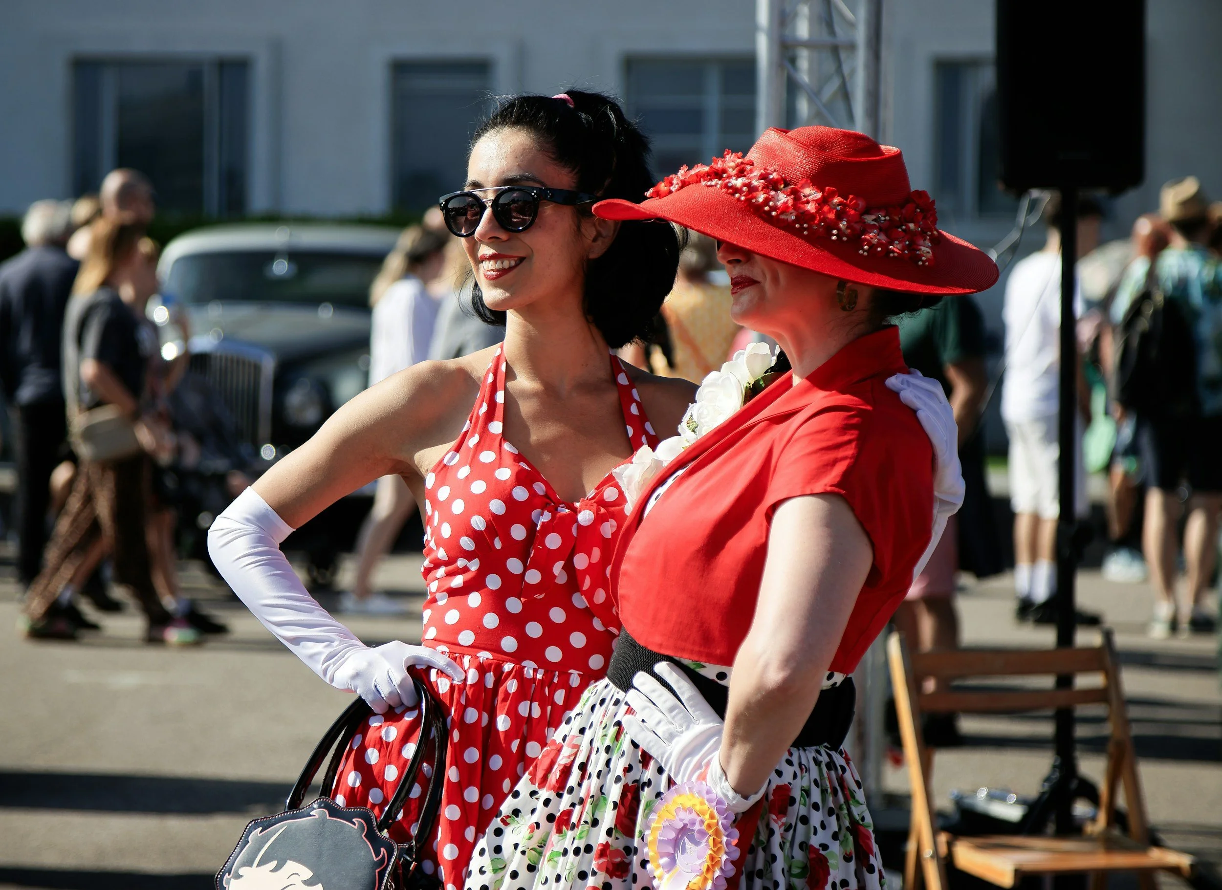 Two women dressed in vintage style clothing with polka dots, wearing wide-brimmed hats, standing outdoors at a daytime event or parade.