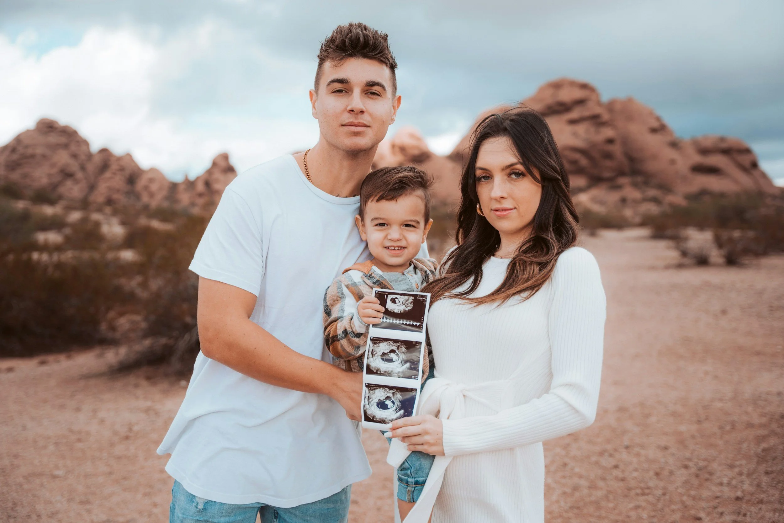 A family of three, including a man, woman, and a young boy, standing outdoors in a desert landscape. The boy is holding ultrasound images, indicating a pregnancy.