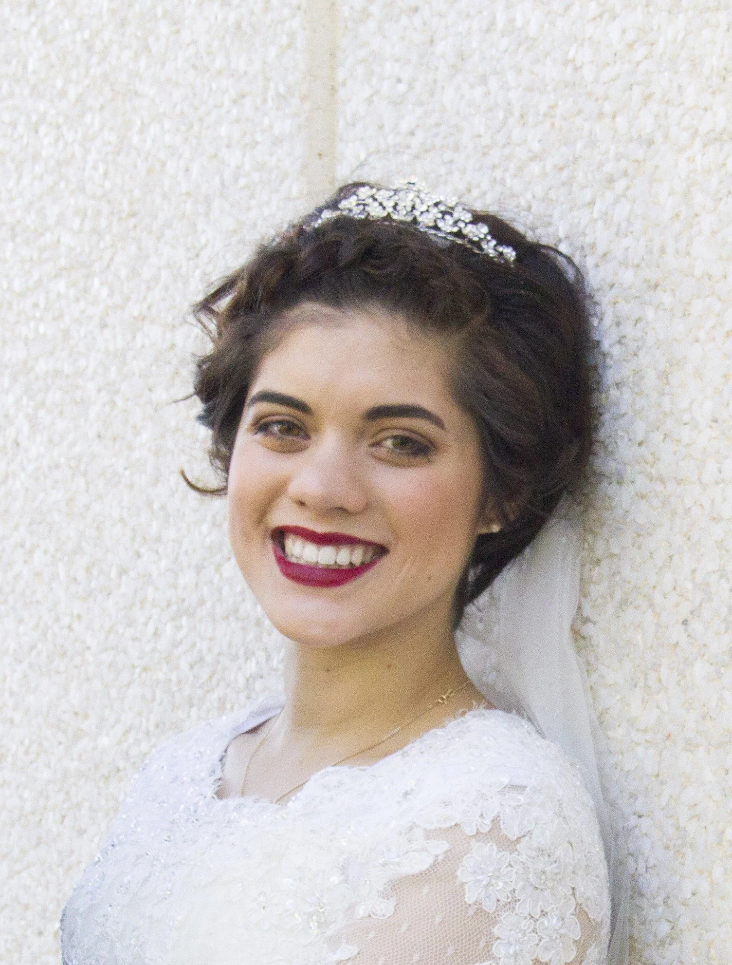 A smiling woman with short, dark hair and a tiara on her head, wearing a white lace wedding dress, standing against a textured beige wall.