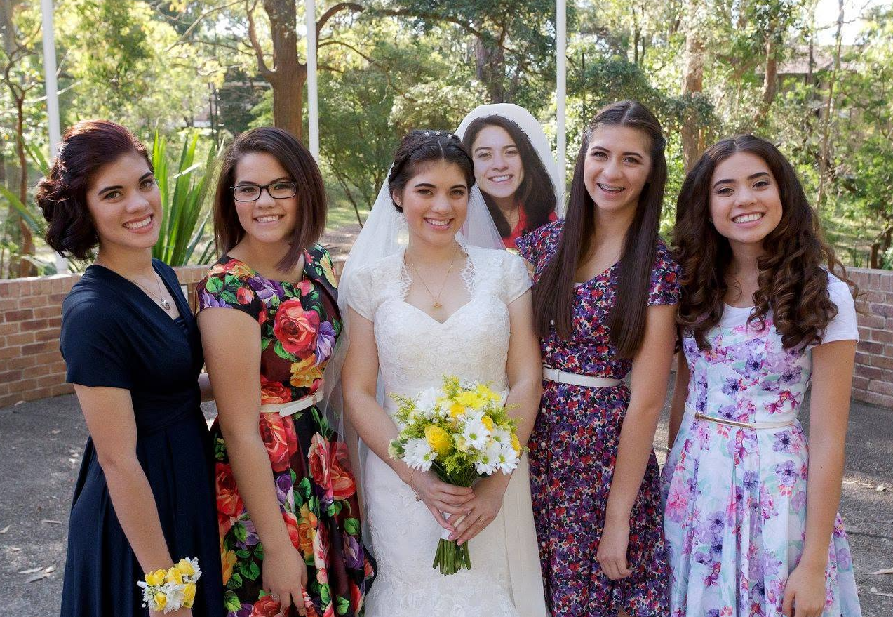 Bride in a white dress holding a bouquet standing with five women in colorful dresses at an outdoor wedding.