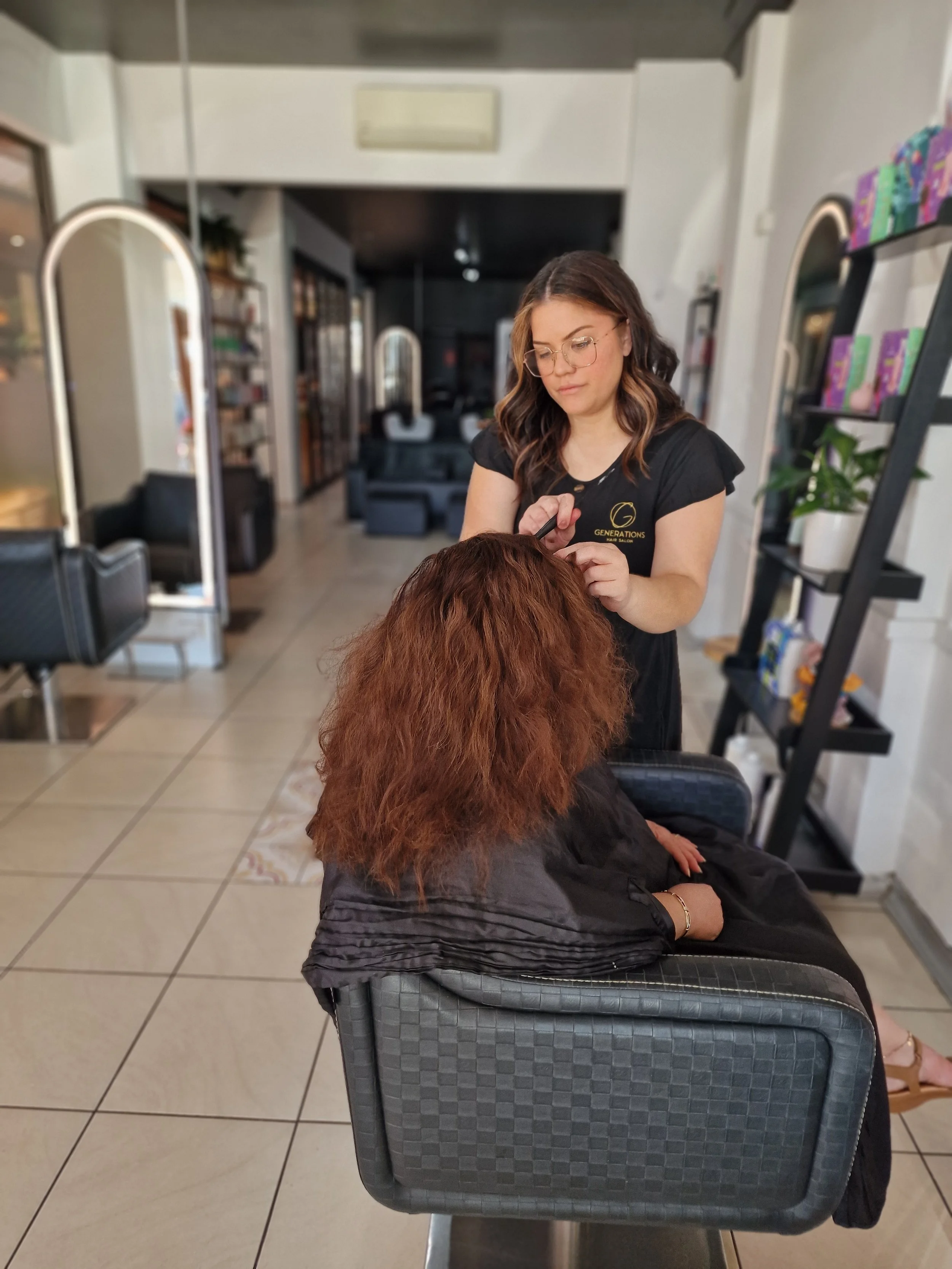 A woman with glasses getting her hair styled in a salon, seated with her arms crossed over her lap, facing away from the camera.