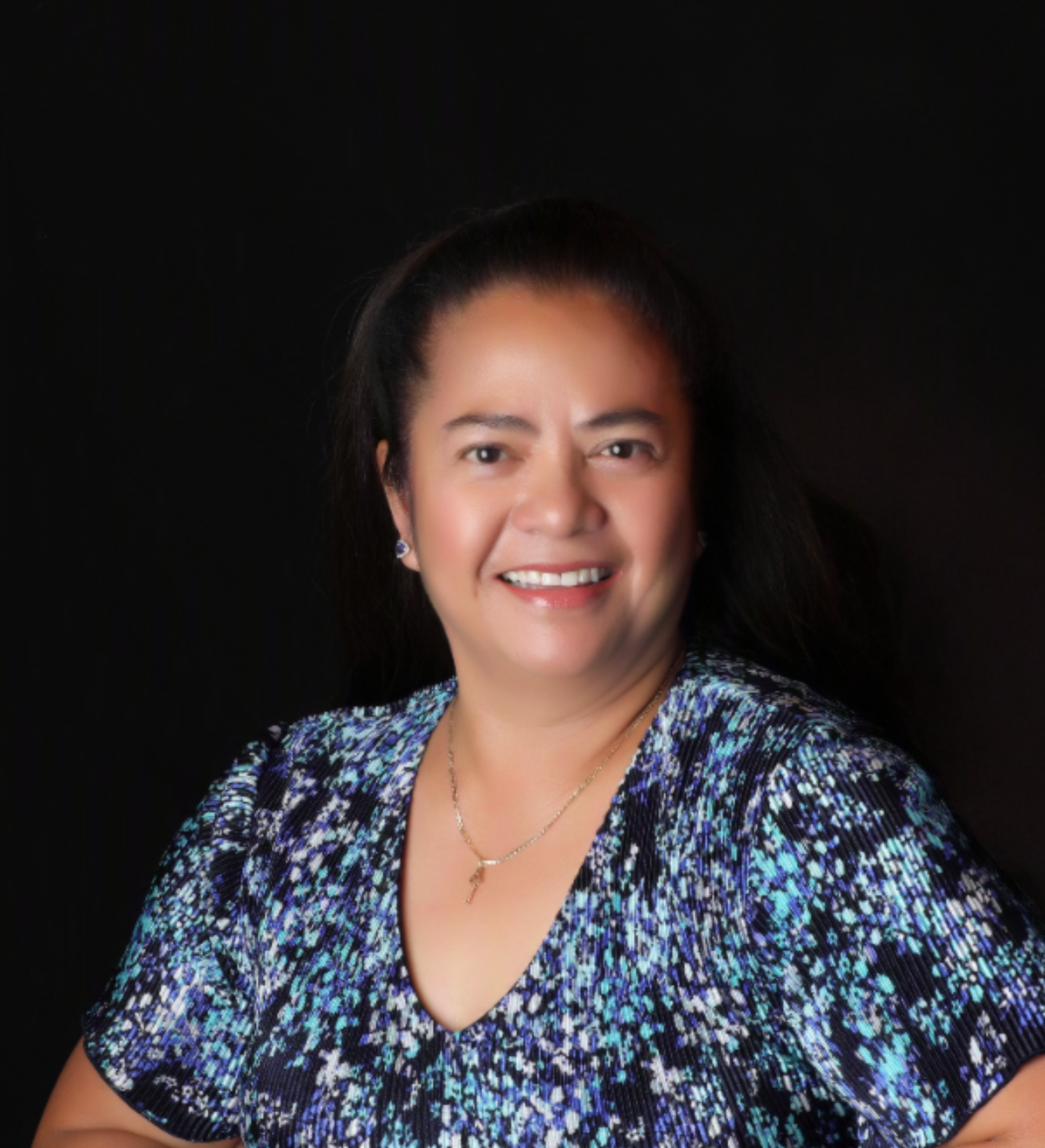 Smiling woman with dark hair wearing a blue and black patterned dress, silver earrings, and a necklace, against a black background.