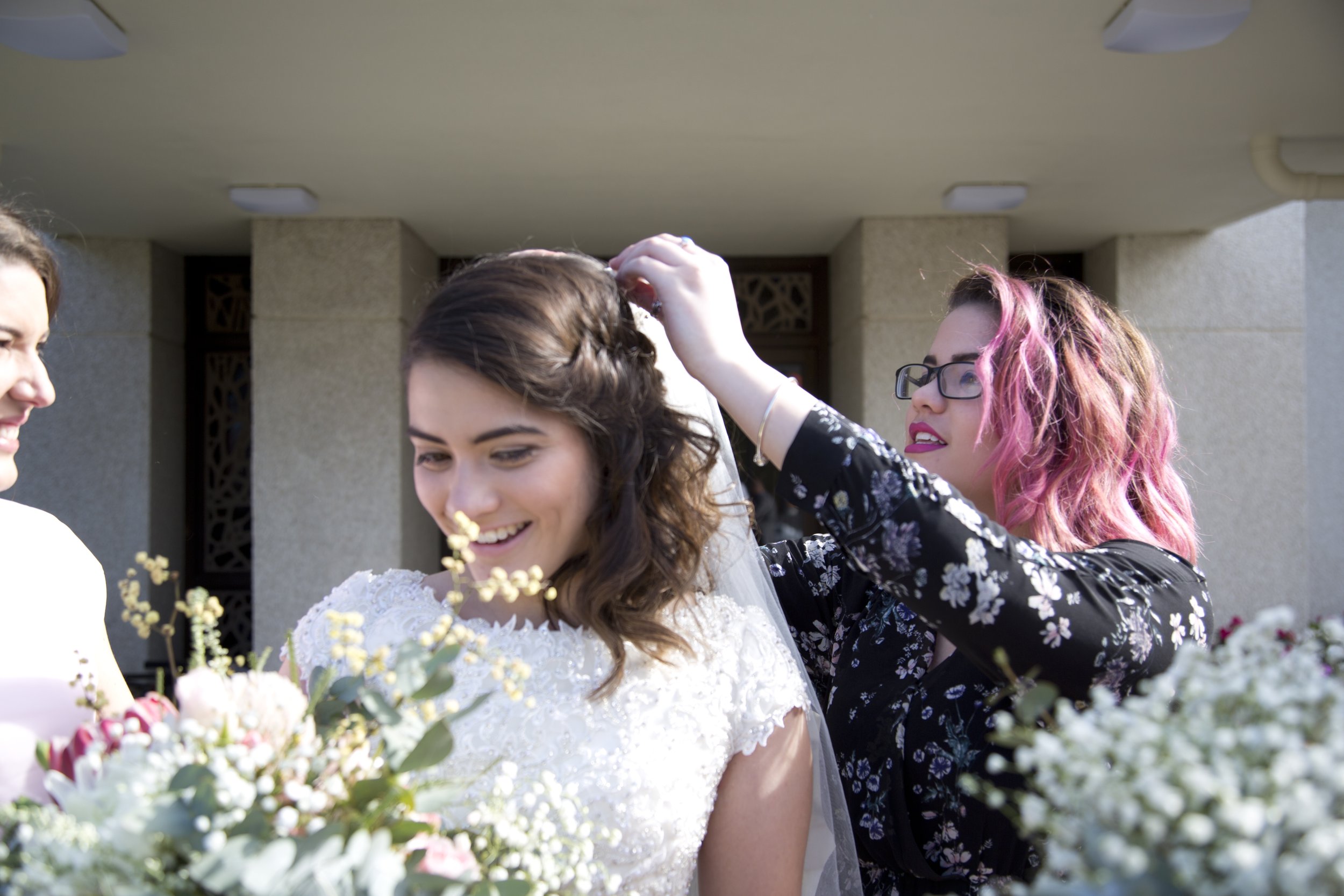 A wedding scene with a bride in a white lace dress smiling, surrounded by flowers, with friends adjusting her veil outside a building with concrete walls and decorative lattice in the background.