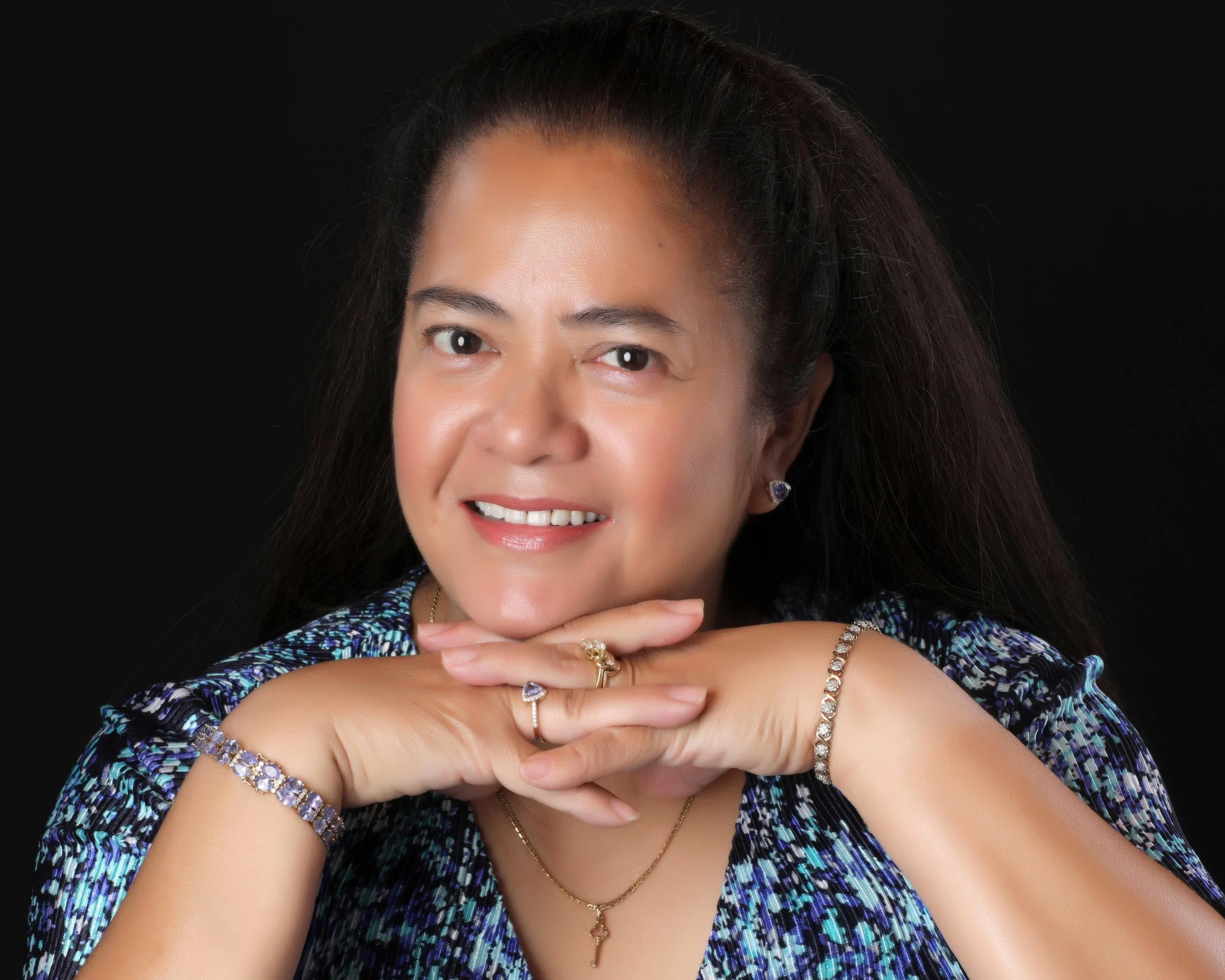 Close-up of a woman with dark, wavy hair, smiling with her chin resting on her hand, wearing multiple jewelry pieces including rings, bracelets, earrings, and layered necklaces, against a black background.