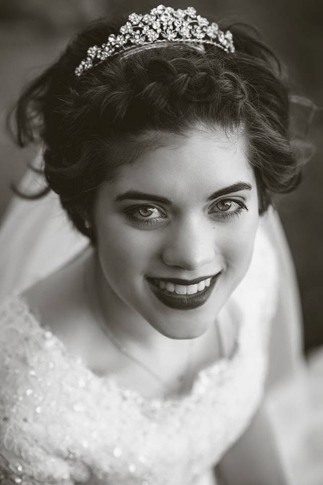 Black and white photo of a smiling bride with dark hair styled in curls, wearing a decorative tiara and a lace wedding dress.