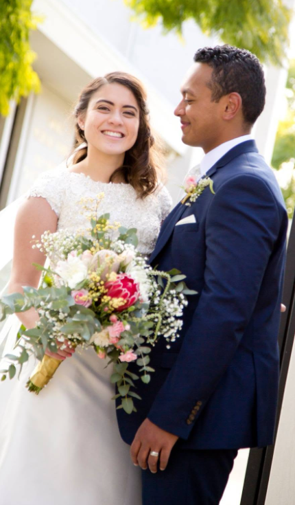 Bride and groom on their wedding day, smiling outdoors, with trees and a house in the background. The bride holds a bouquet of flowers.