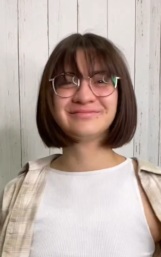 Young woman with short brown hair, glasses, and a white tank top, standing in front of a white wooden wall.