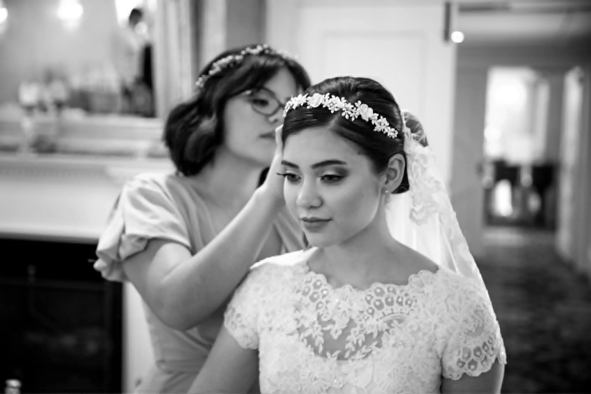 A woman in a lace wedding dress with a floral headband is being assisted by another woman, possibly a makeup artist or stylist, in a preparation area.
