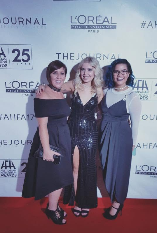 Three women posing on red carpet at an awards event with a backdrop displaying logos and event information.