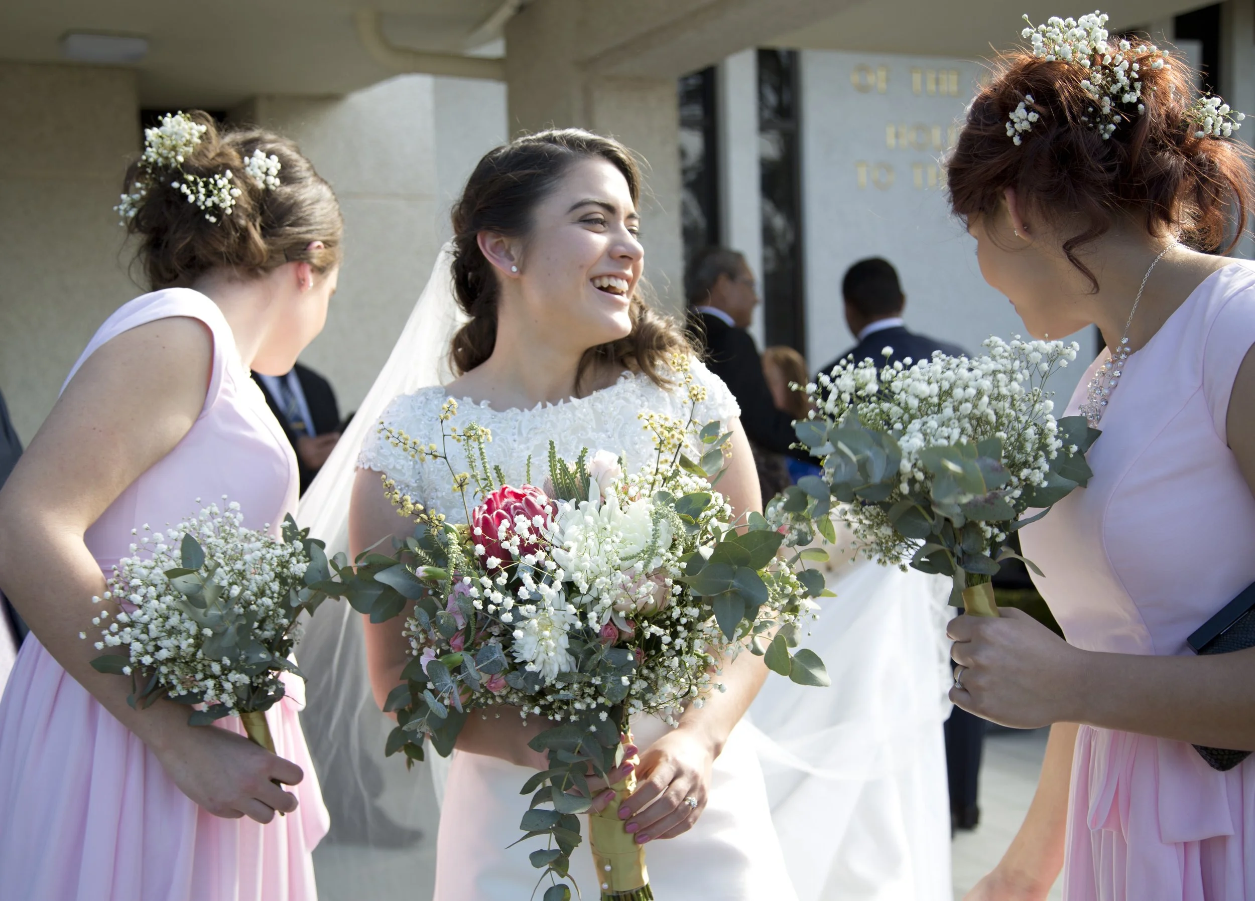 The image shows a bride in a white wedding dress smiling and holding a bouquet of flowers, while being surrounded by bridesmaids in pink dresses, also holding bouquets. They are outdoors in front of a building with some people in the background.