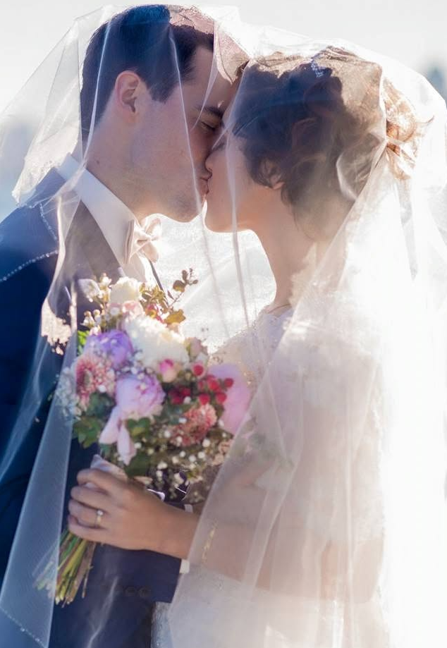 A bride and groom kiss under a sheer veil, with the bride holding a colorful bouquet of flowers.