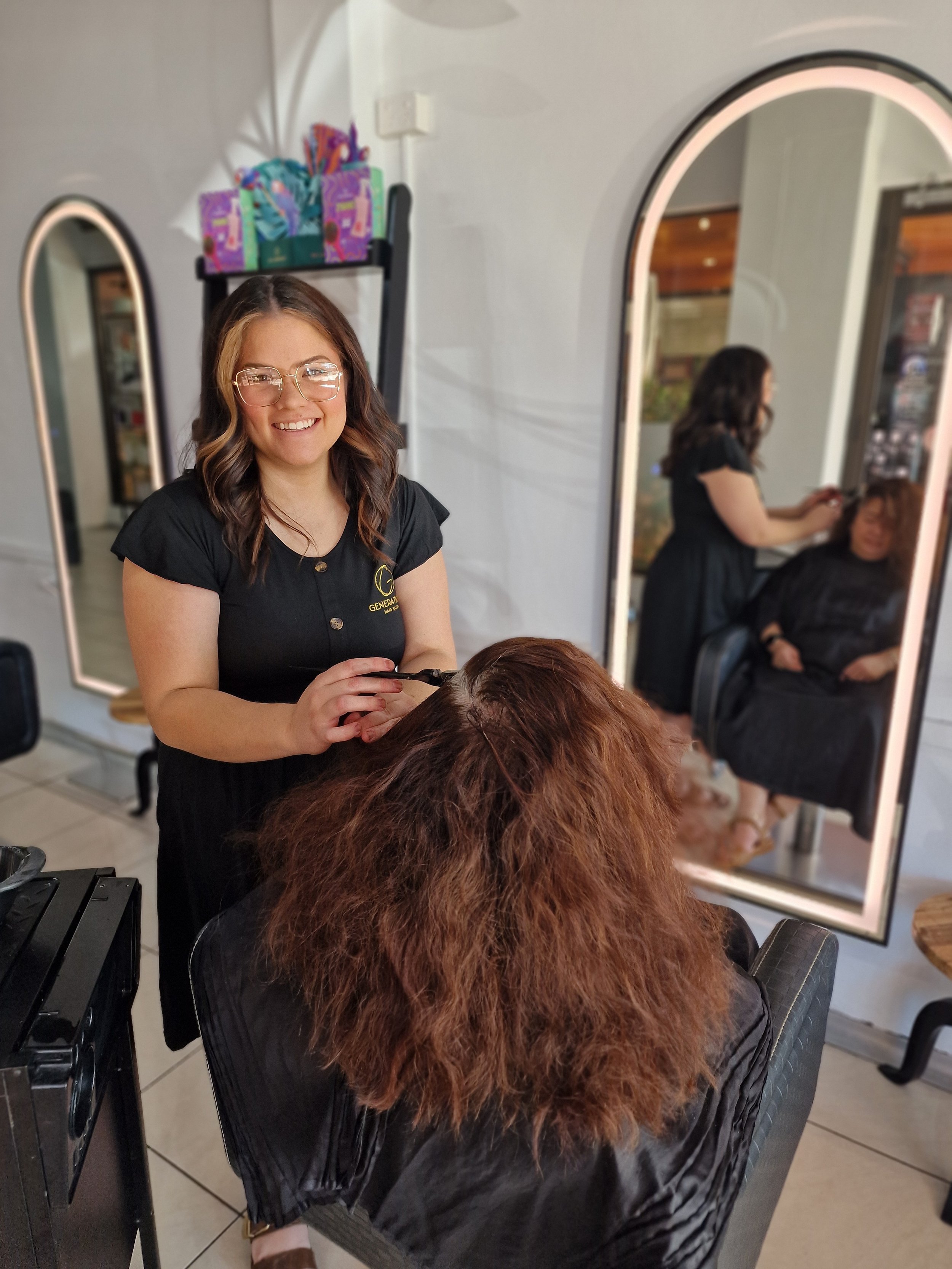 A hairstylist smiling and working on a woman's hair in a salon. The woman is seated with her back to the camera, and her hair is reddish-brown and voluminous. There is a mirror reflecting the scene.