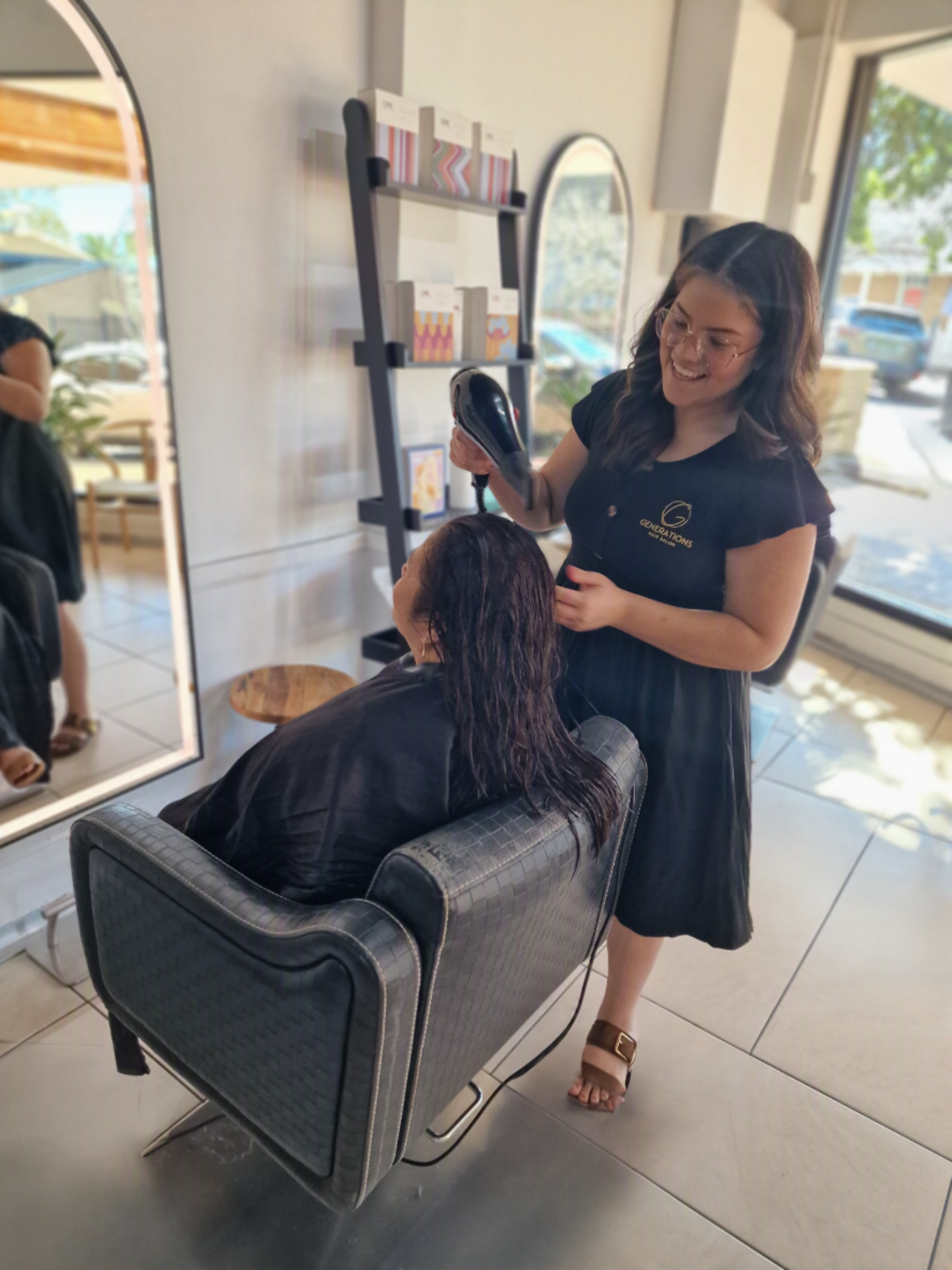A woman in a black dress, wearing glasses and sandals, standing next to a seated person with long, wet hair inside a salon. The woman is holding a hairdryer and smiling.
