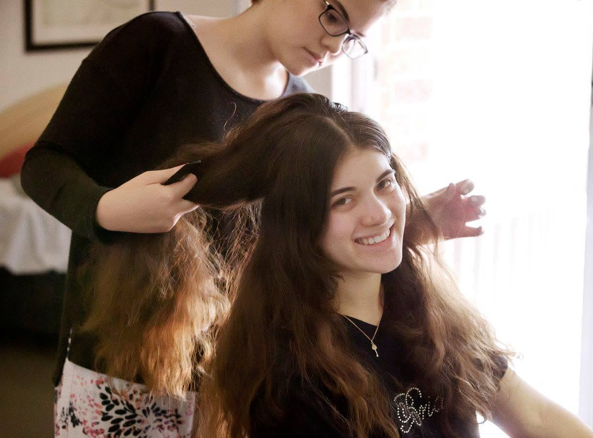A woman with long wavy brown hair smiling in a salon chair while a stylist with glasses brushes her hair.