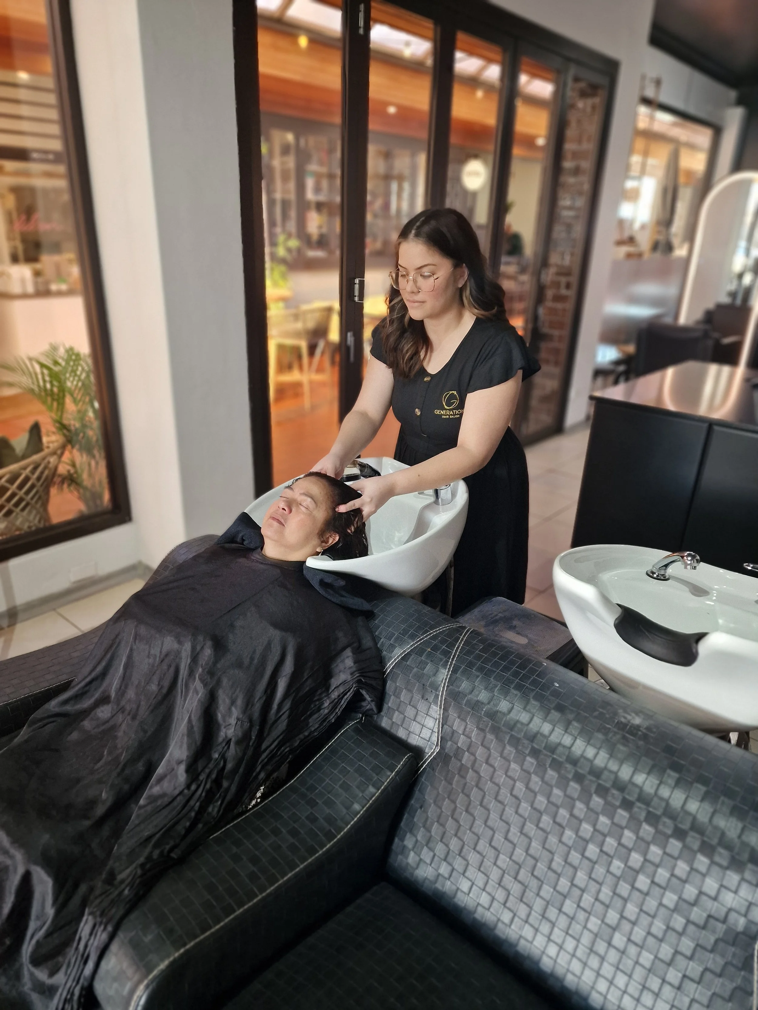 A woman receiving a hair wash at a salon, with her head tilted back into a white sink as a stylist rinses her hair.