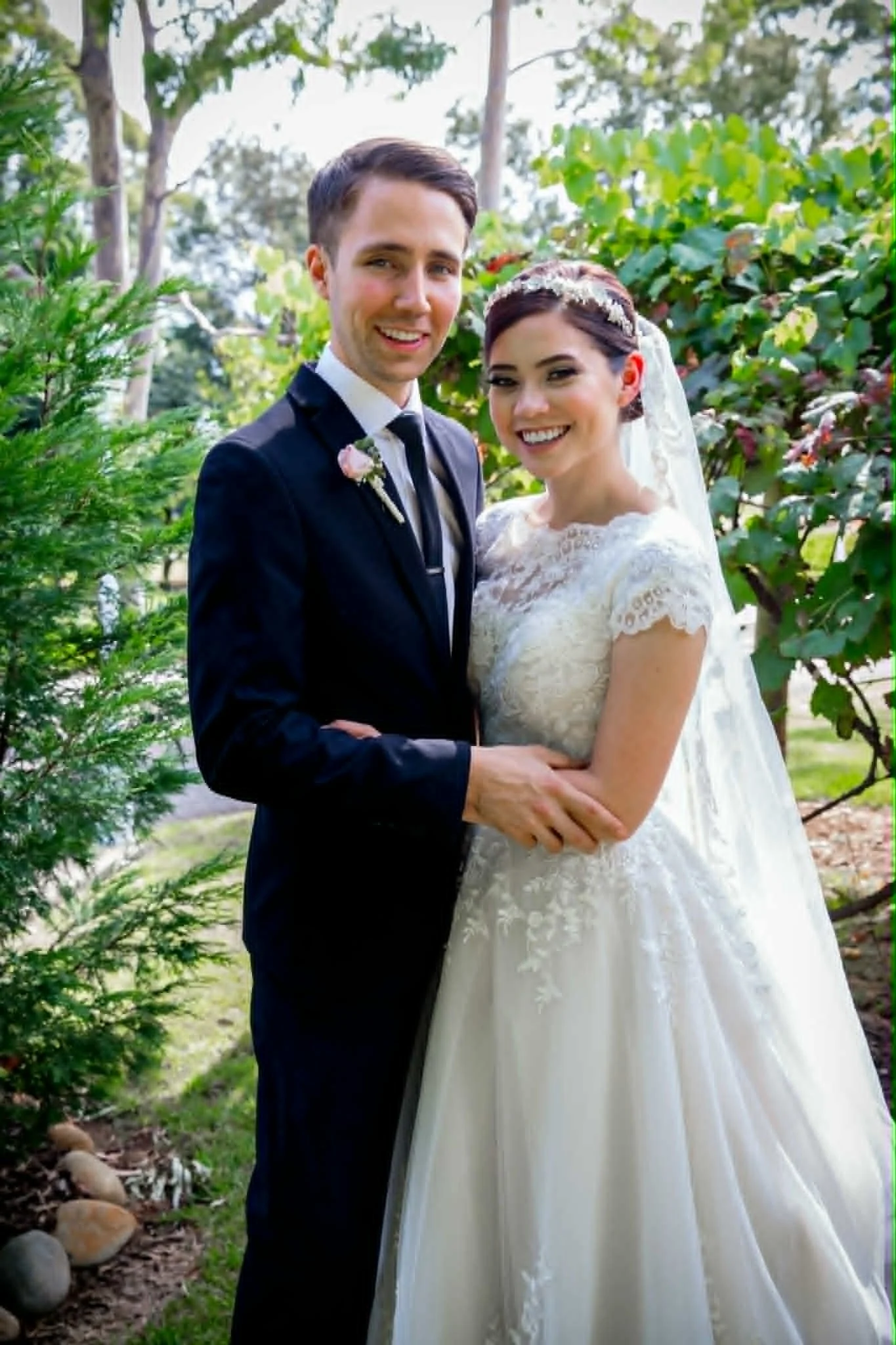 A happy bride and groom standing outdoors in a garden, smiling, with the bride in a white lace wedding dress and the groom in a dark suit with a boutonniere, surrounded by greenery and trees.