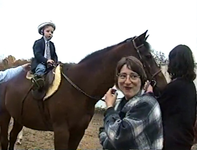 Child riding a horse with two women beside them outdoors on a cloudy day.