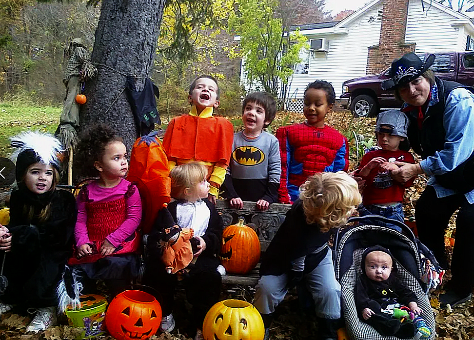 Children dressed in Halloween costumes gathered outdoors with a woman, pumpkins, and autumn leaves, celebrating Halloween.