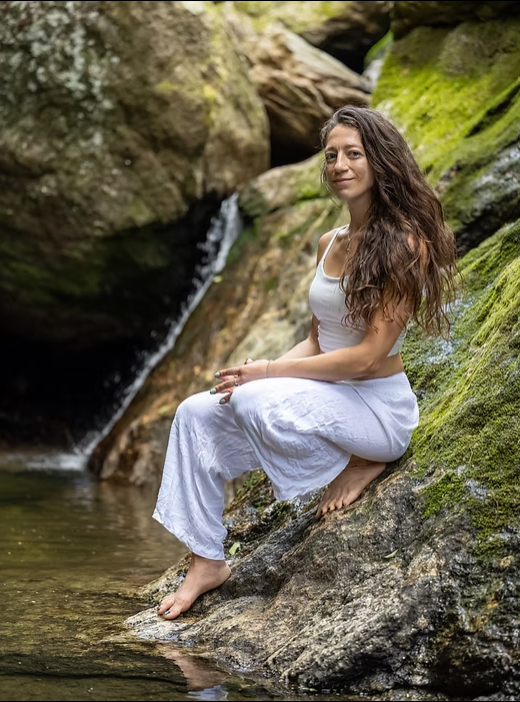 A woman sitting on moss-covered rocks at a creek, wearing a white dress, with long, wavy brown hair and a peaceful expression, surrounded by greenery and large rocks. It is a photo of the instructor.