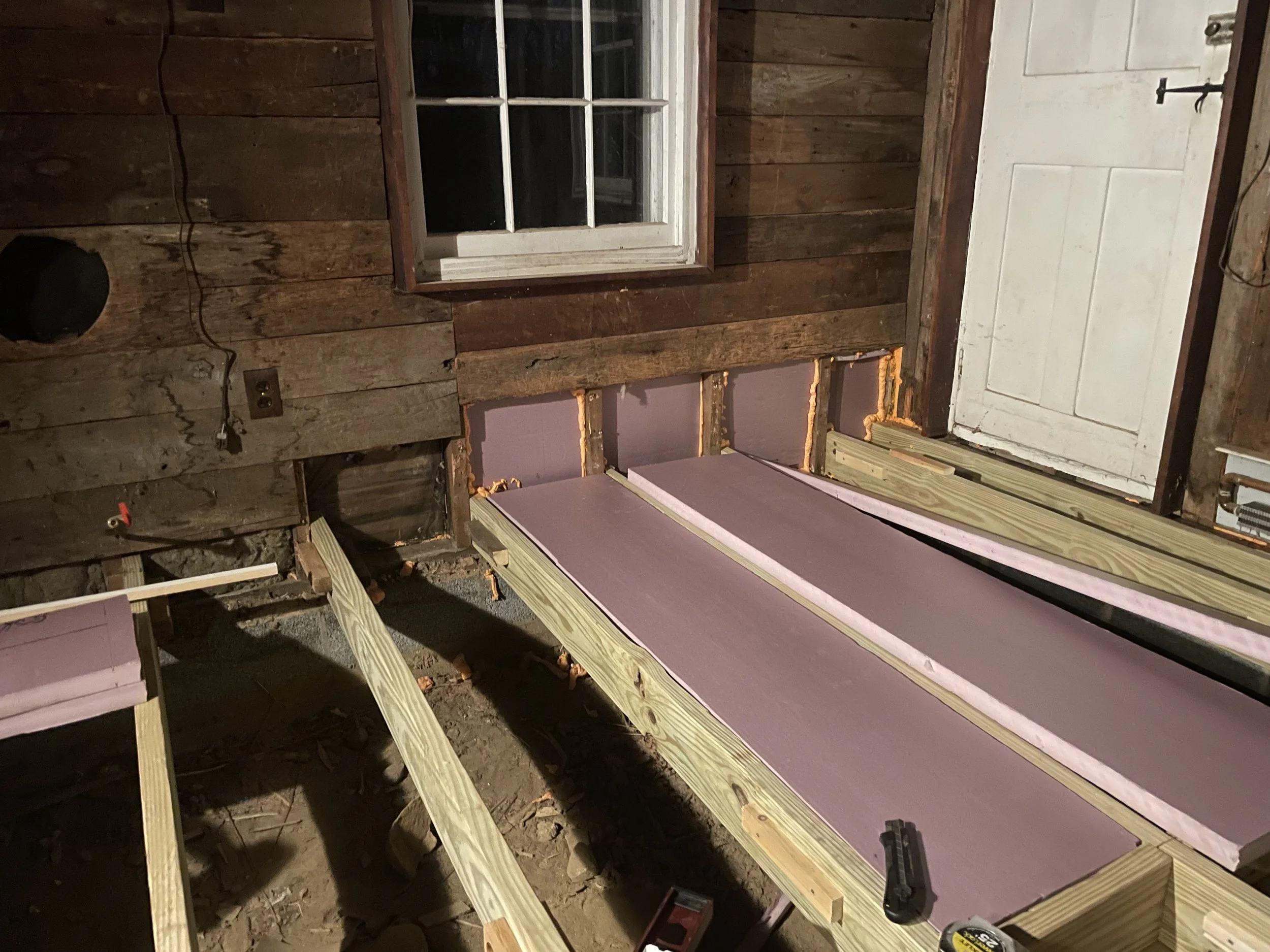 A room under construction with wooden framing, purple drywall panels, a window on the wall, and construction tools on the floor.