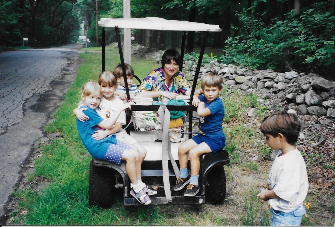 A group of children and an adult woman sitting and standing on a golf cart parked on the side of a paved road, surrounded by green trees and rocks in a forested area.