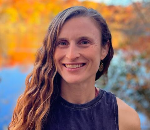 A woman with long, wavy hair smiling outdoors with a blurred autumn background. It is a photo of the instructor.