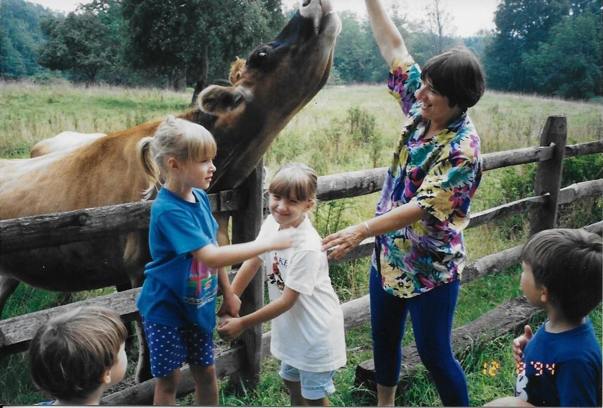 A woman and children at a farm, with a horse reaching over a wooden fence to interact with them.