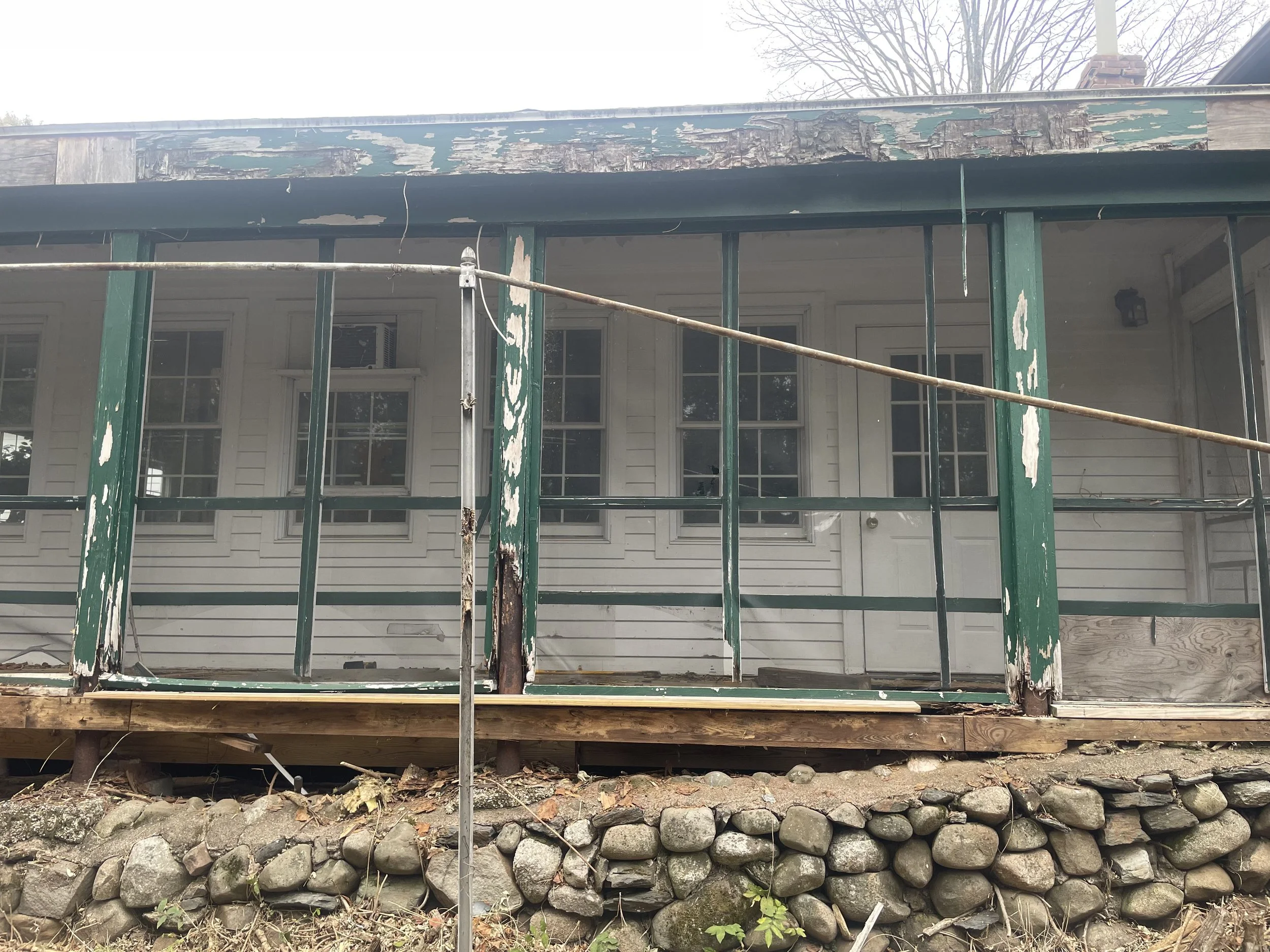 A house with a porch and window air conditioning unit, undergoing renovations with green and peeling paint siding, scaffolding in front, and a rocks foundation.