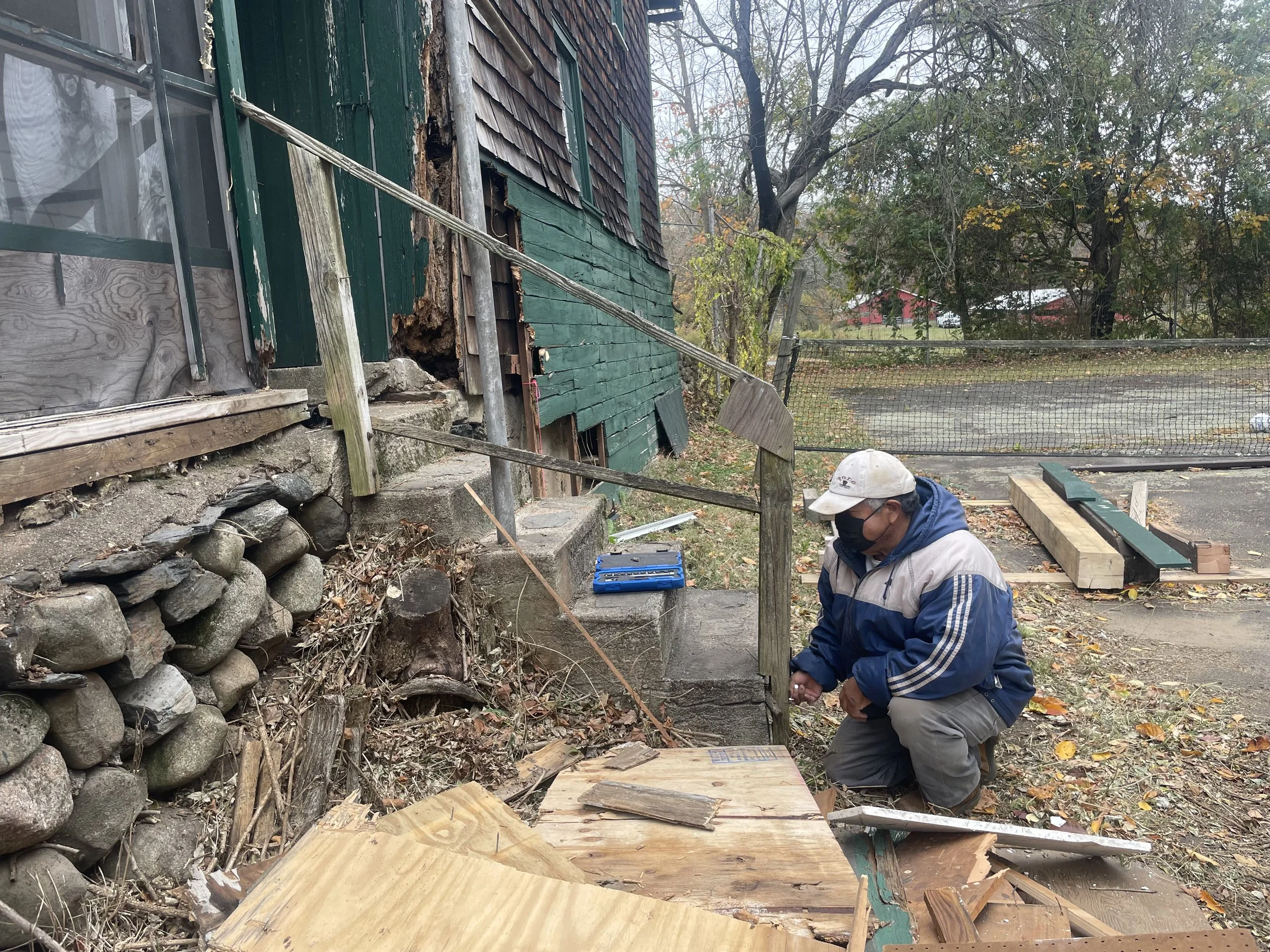 A person wearing a cap and face mask crouches down working on some wood planks in front of a damaged house with a rotting exterior, a stone foundation, and a fallen wooden post. There are trees and a chain-link fence in the background.