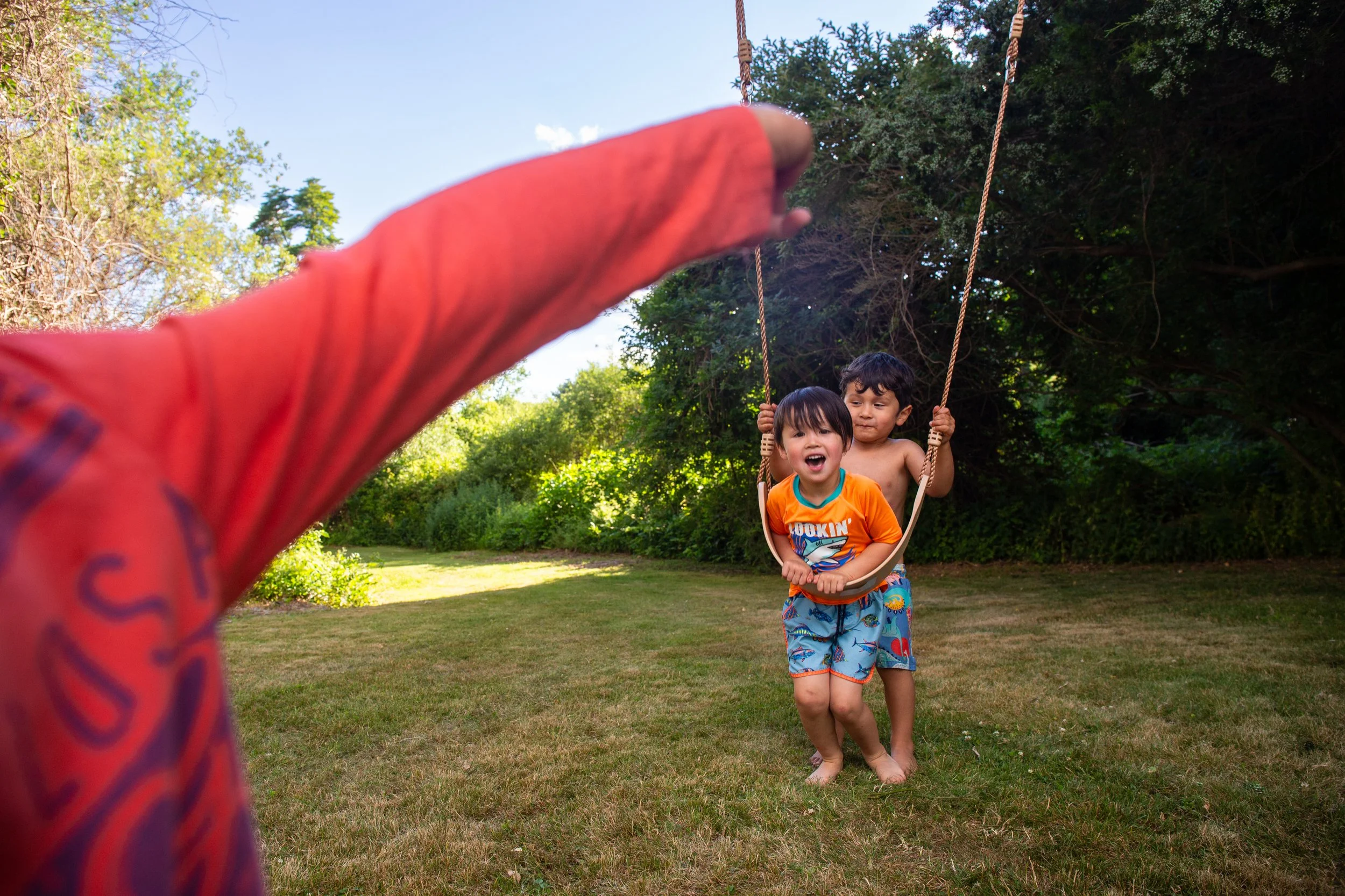 Two children, a boy and a girl, playing on a swing in a grassy outdoor area with trees in the background. The girl is smiling, and the boy is looking on as the swing extends backward, while an out-of-focus arm in red sleeves is reaching toward the children from the foreground.