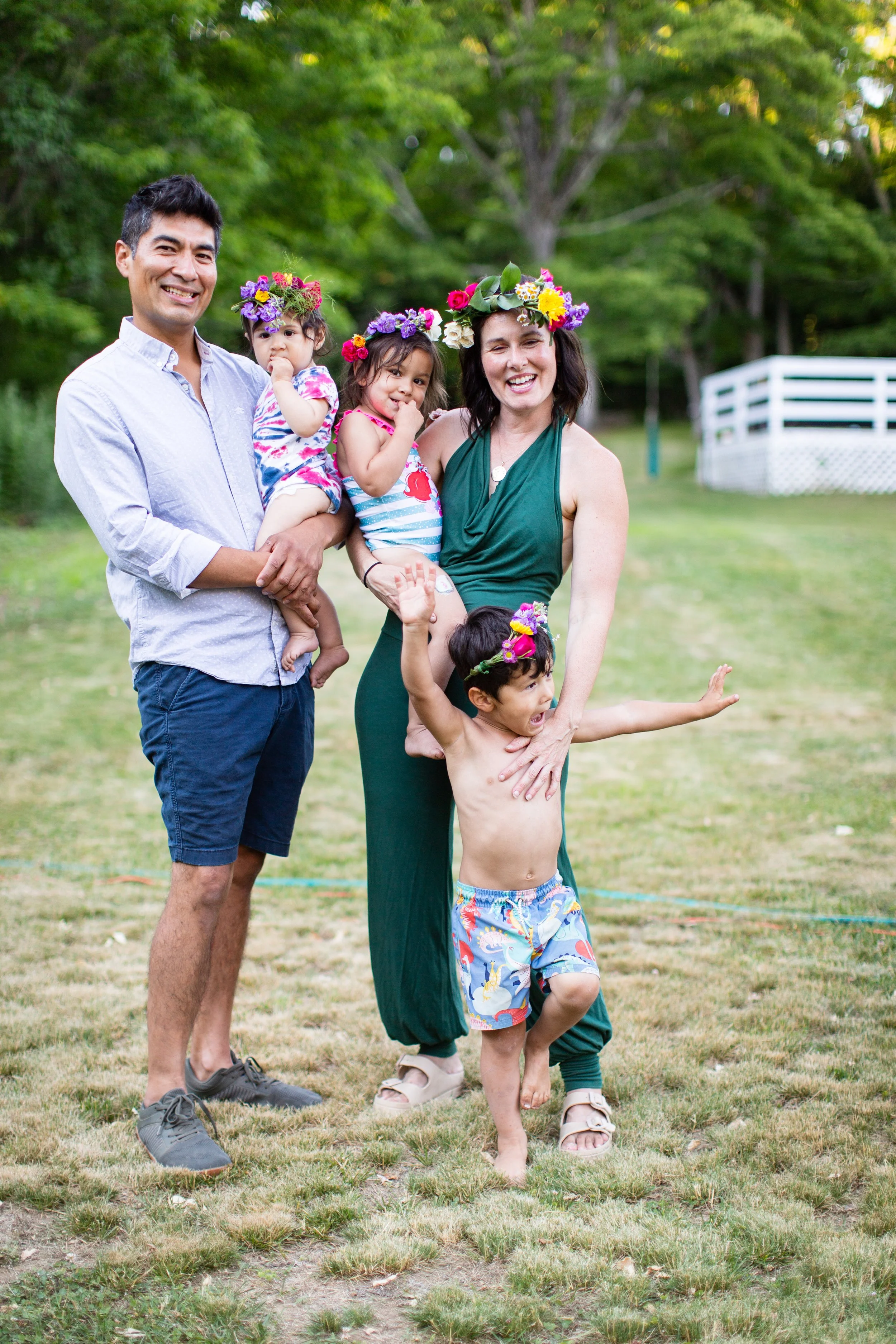 A happy family of five standing on a grassy field outdoors, wearing flower crowns, with a green background of trees and a white fence. The parents are smiling, and the children, barefoot or in sandals, are playing.