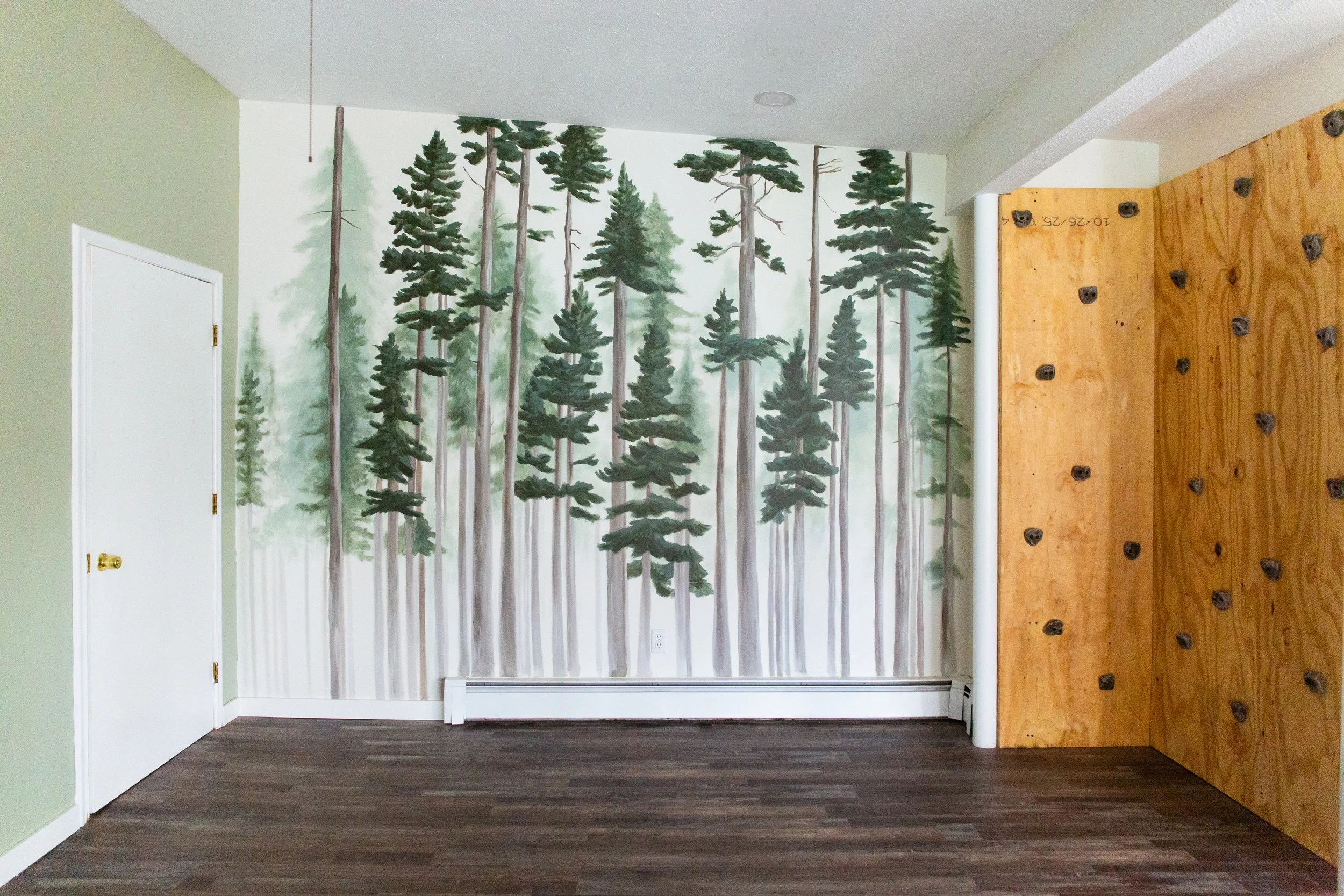A room with a wall painted with a forest scene featuring tall pine trees and green foliage. The room has a white door with a gold handle on the left, a white baseboard heater beneath the mural, light green painted walls, and dark wood flooring. There is also a wooden climbing wall on the right with black holds.