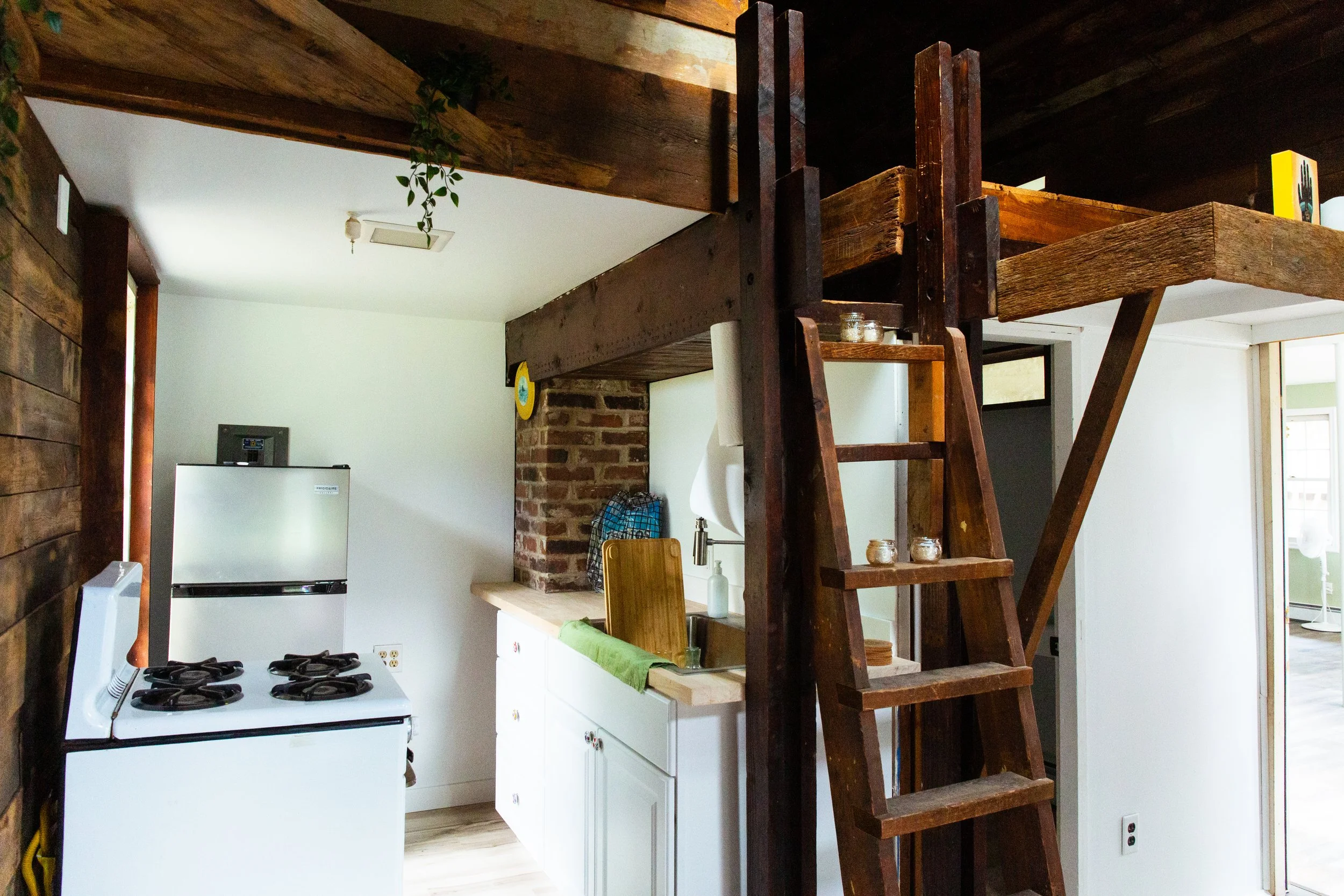 Interior of a cozy kitchen with wooden beams, white cabinets, a gas stove, and a brick accent wall, viewed from an adjacent room with wooden elements.